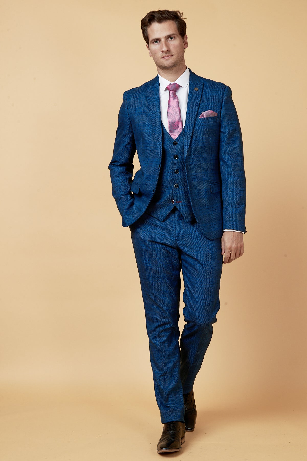 A man in the marcdarcy JERRY Blue Check Suit with a single breasted waistcoat, paired with a white shirt and pink paisley tie, stands with his hand in his pocket.