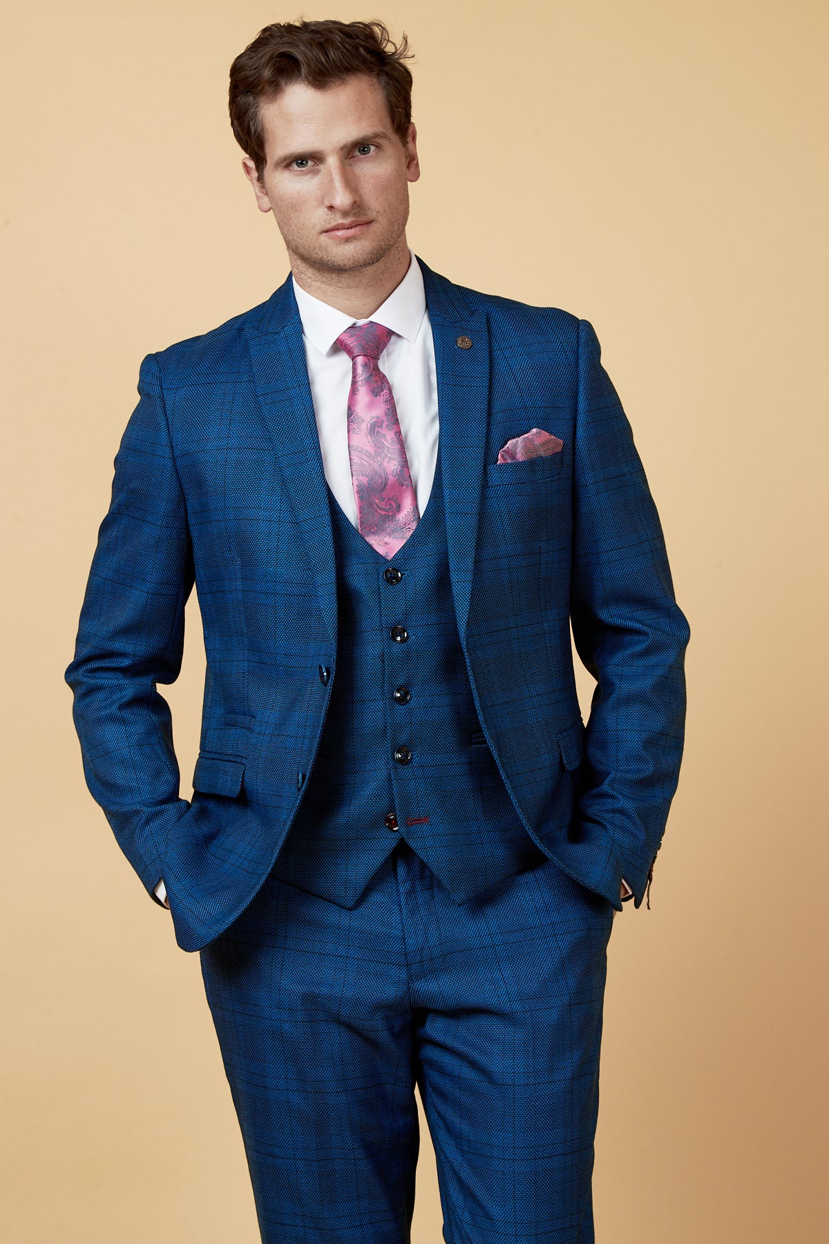 A man wears the marcdarcy JERRY Blue Check Suit with a single breasted waistcoat, paired with a pink patterned tie and matching pocket square, standing against a beige background with hands in his pockets.