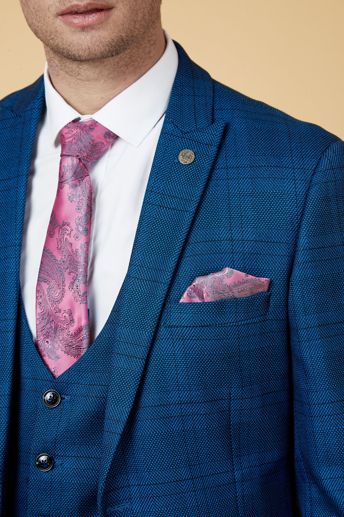 A man wears the marcdarcy JERRY Blue Check Suit with a single breasted waistcoat, paired with a white dress shirt and pink paisley tie, shown from shoulders to mid-chest against a beige background.