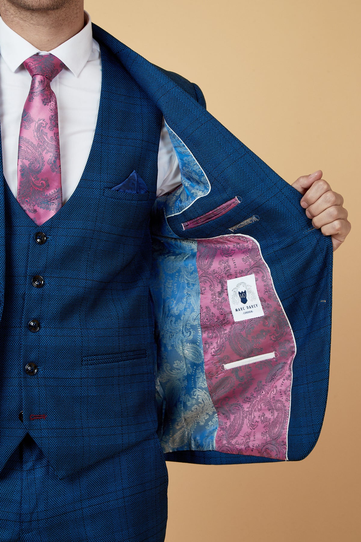 A man in a slim fit JERRY - Blue Check Blazer by marcdarcy with a pink paisley tie holds open his jacket, showing a vibrant blue and pink paisley lining, interior pockets, and the Bake Angel label.