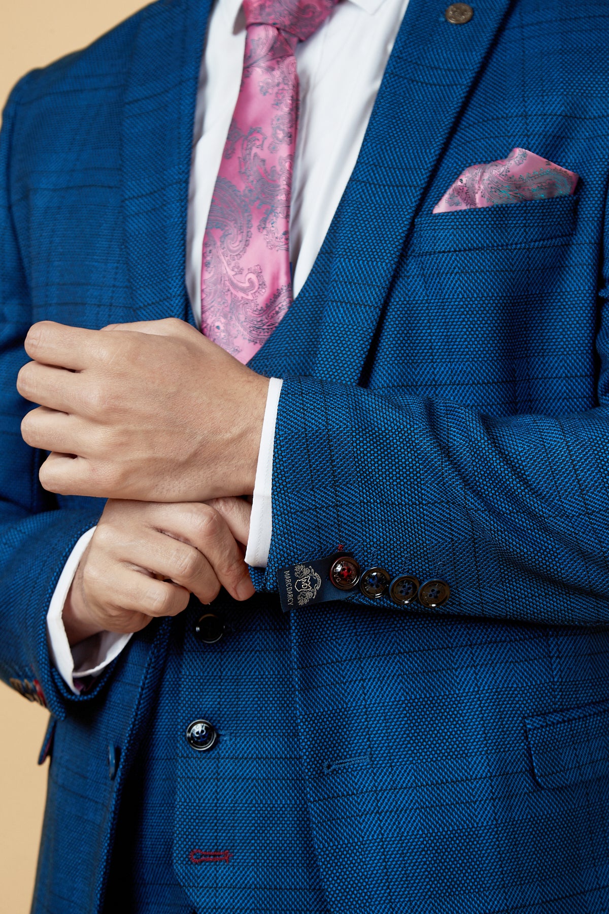 A man wears the marcdarcy JERRY - Blue Check Blazer, paired with a white shirt and pink paisley tie, adjusting his cuff. A pink pocket square accents his look. His face is not visible.