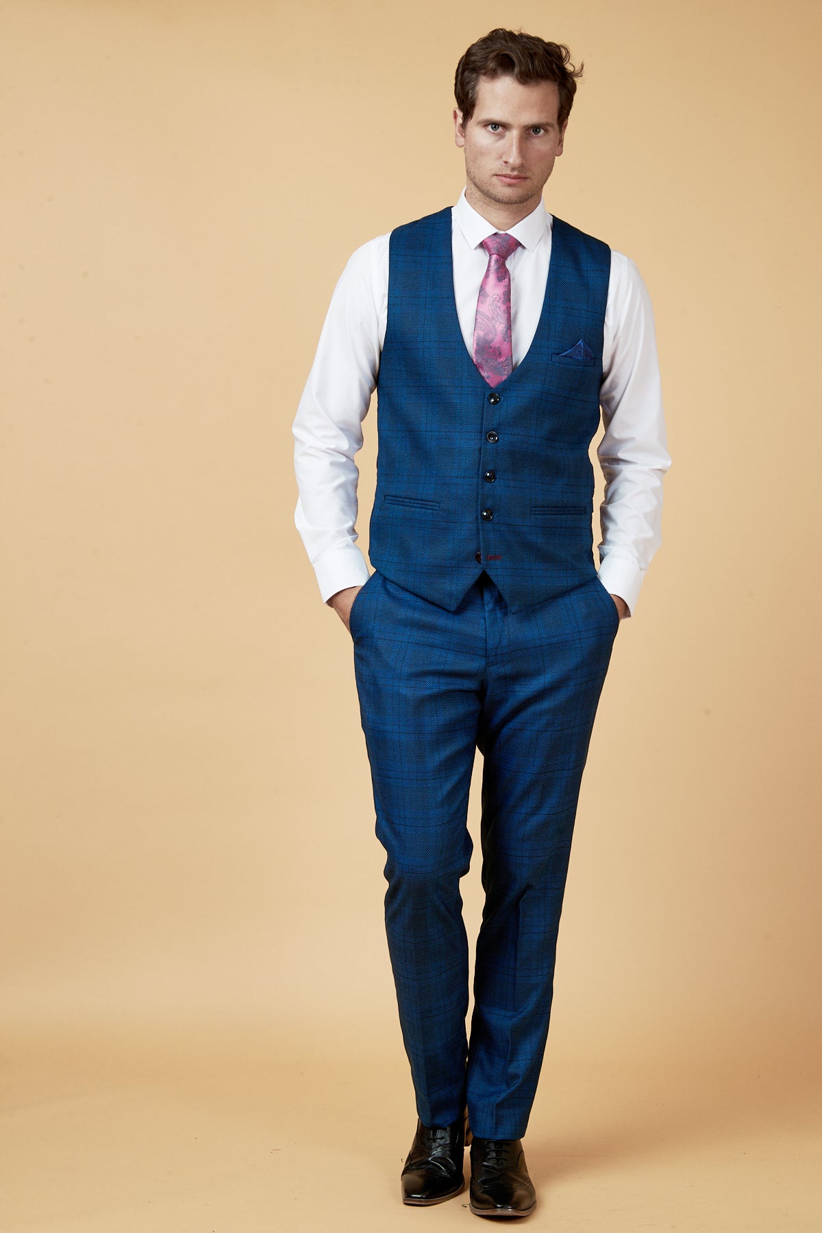 A man poses against a beige background wearing the marcdarcy JERRY - Blue Check Single Breasted Waistcoat, paired with a white shirt and pink patterned tie. He stands with his hands in his pockets, looking slightly to the side.