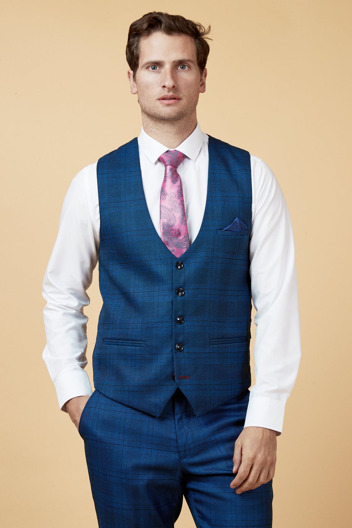 A man wears the marcdarcy JERRY Blue Check Single Breasted Waistcoat with matching trousers, a white shirt, and a pink patterned tie, standing against a beige backdrop with one hand in his pocket.