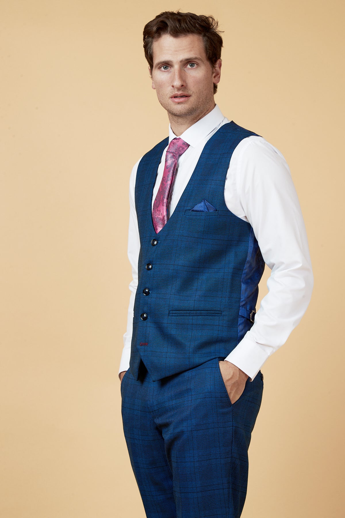 A man wearing the marcdarcy JERRY blue check single breasted waistcoat, pink patterned tie, and white shirt stands with his hand in his pocket against a beige background.
