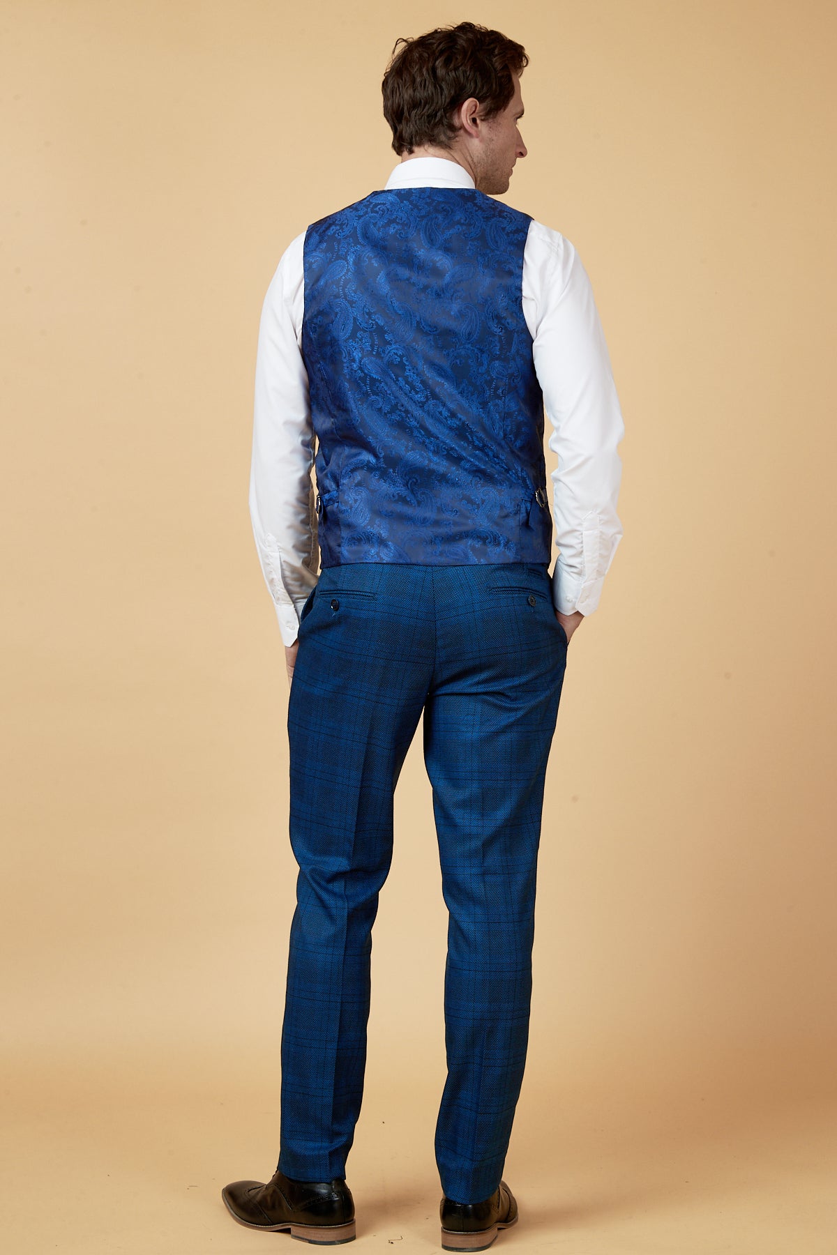 A man stands facing away in a marcdarcy JERRY Blue Check Single Breasted Waistcoat, slim blue plaid dress pants, white long-sleeve shirt, and brown dress shoes against a beige background.