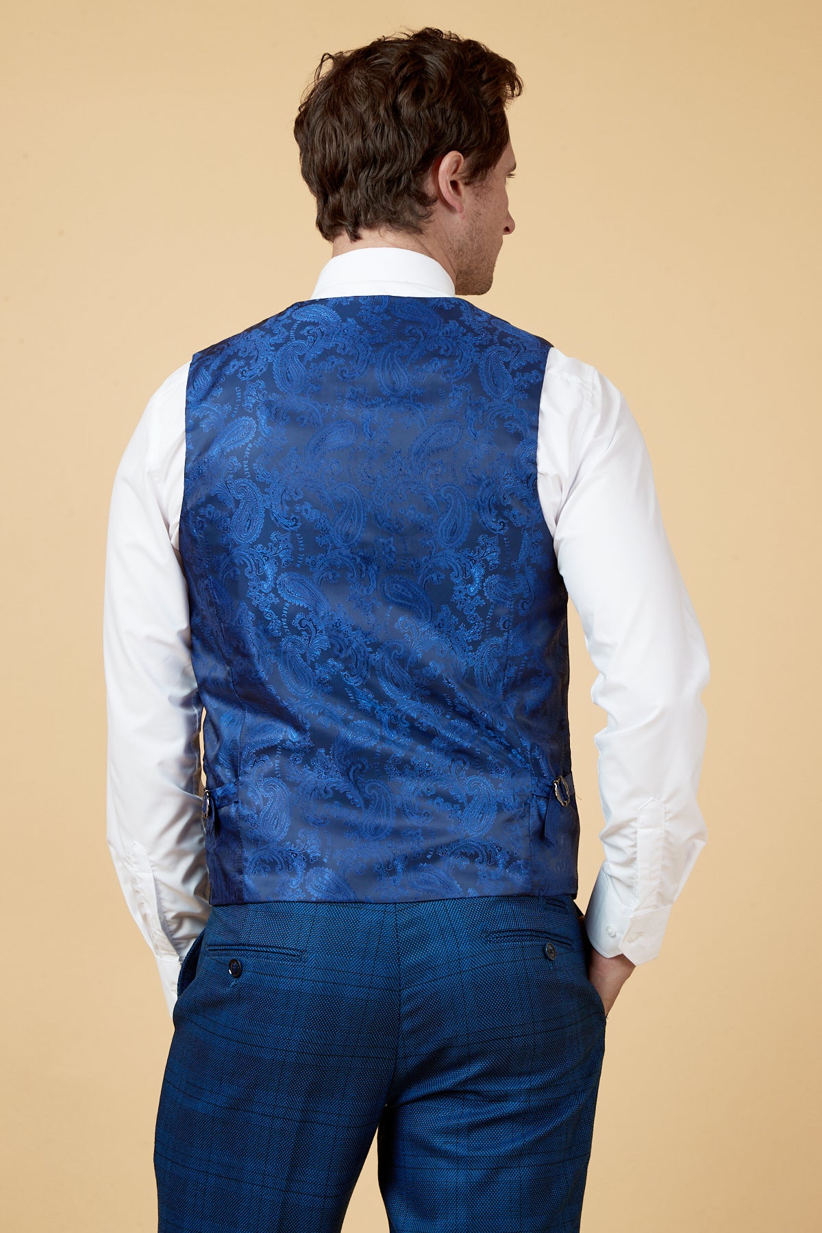 A man stands facing away in a slim white dress shirt and the marcdarcy JERRY Blue Check Single Breasted Waistcoat, paired with blue plaid trousers, set against a beige background.