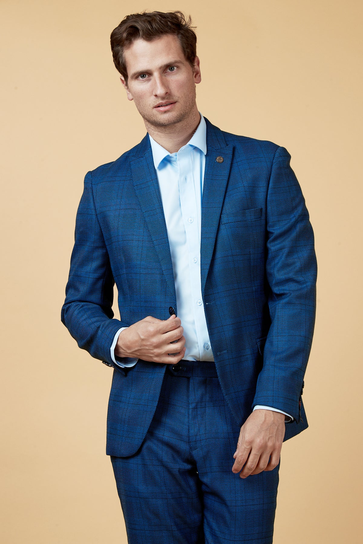 A man wears the marcdarcy JERRY - Blue Check Blazer over a light blue shirt, standing against a beige background facing the camera with a neutral expression, one hand adjusting his jacket.