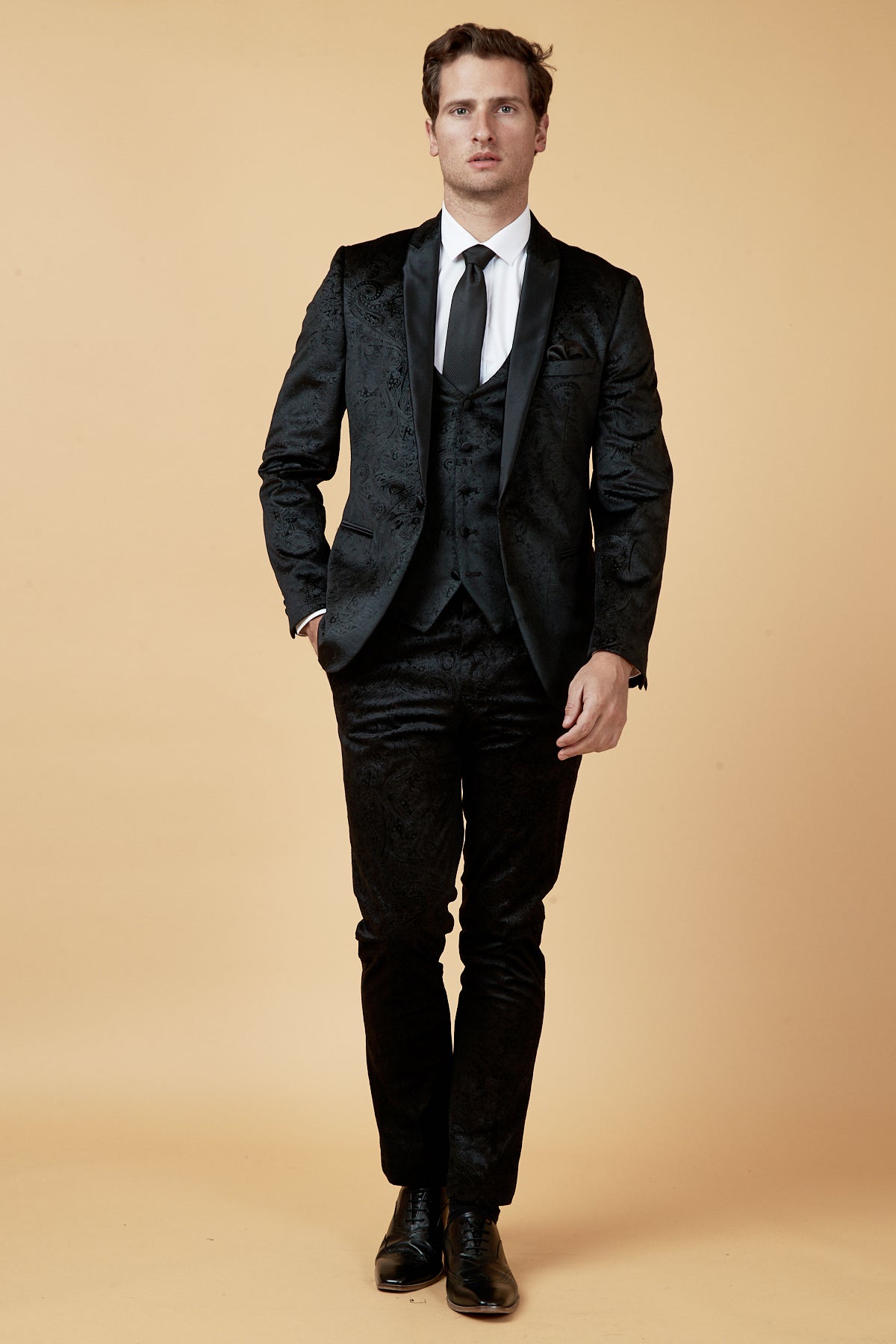 A man faces forward against a beige background, wearing the Marc Darcy SIMON Black Velvet Jacquard Three Piece Suit with satin lapel, white shirt, black tie, and black dress shoes—ideal for sophisticated events.