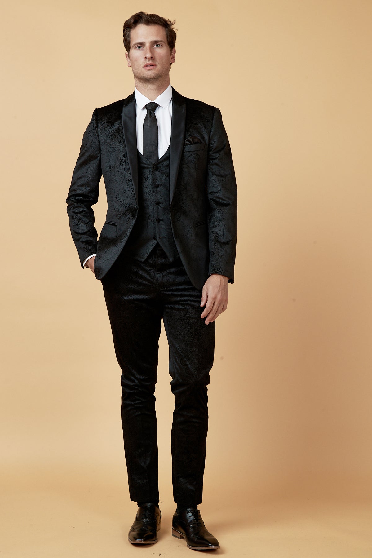 A man with short brown hair and a neutral expression stands against a beige background wearing the Marc Darcy SIMON Black Velvet Jacquard Three Piece Suit, featuring a satin shawl collar, white shirt, black tie, and shoes.