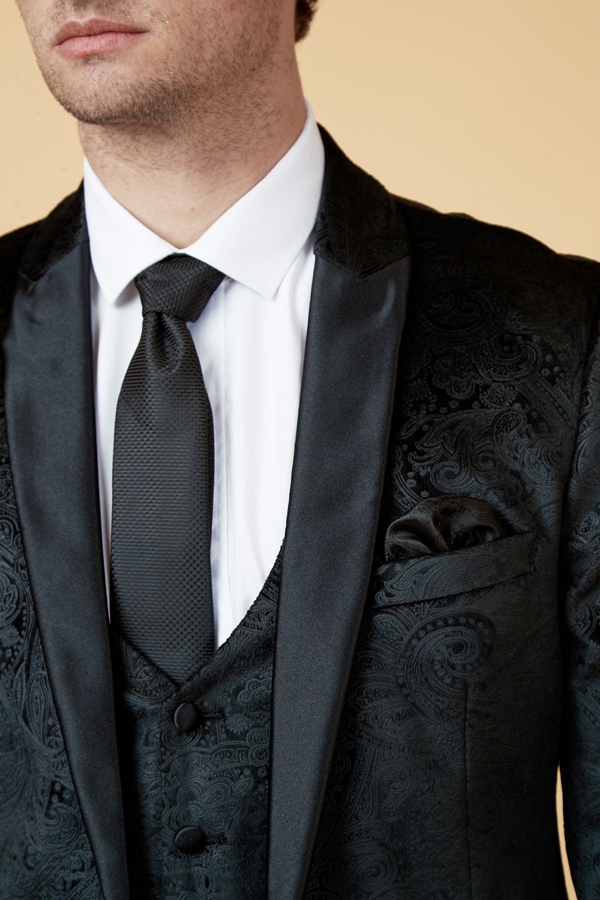 A close-up of a man in the Marc Darcy SIMON Black Velvet Jacquard Three Piece Suit with satin shawl collar, white shirt, black tie, and matching pocket square. The background is solid light beige; his face is partially visible.