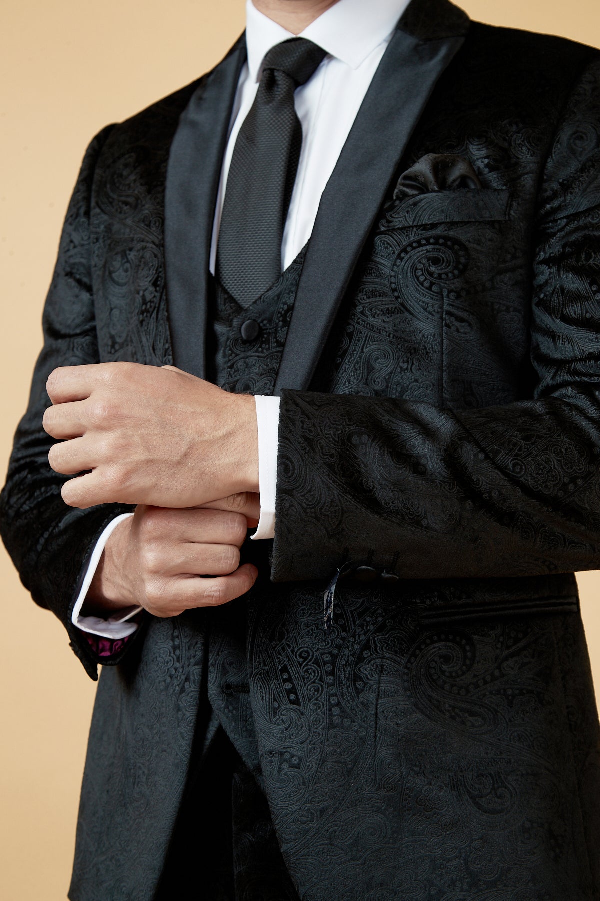 A man in the Marc Darcy SIMON black velvet jacquard three piece suit with a satin shawl collar adjusts his cuff against a light beige backdrop, his face partially out of frame.