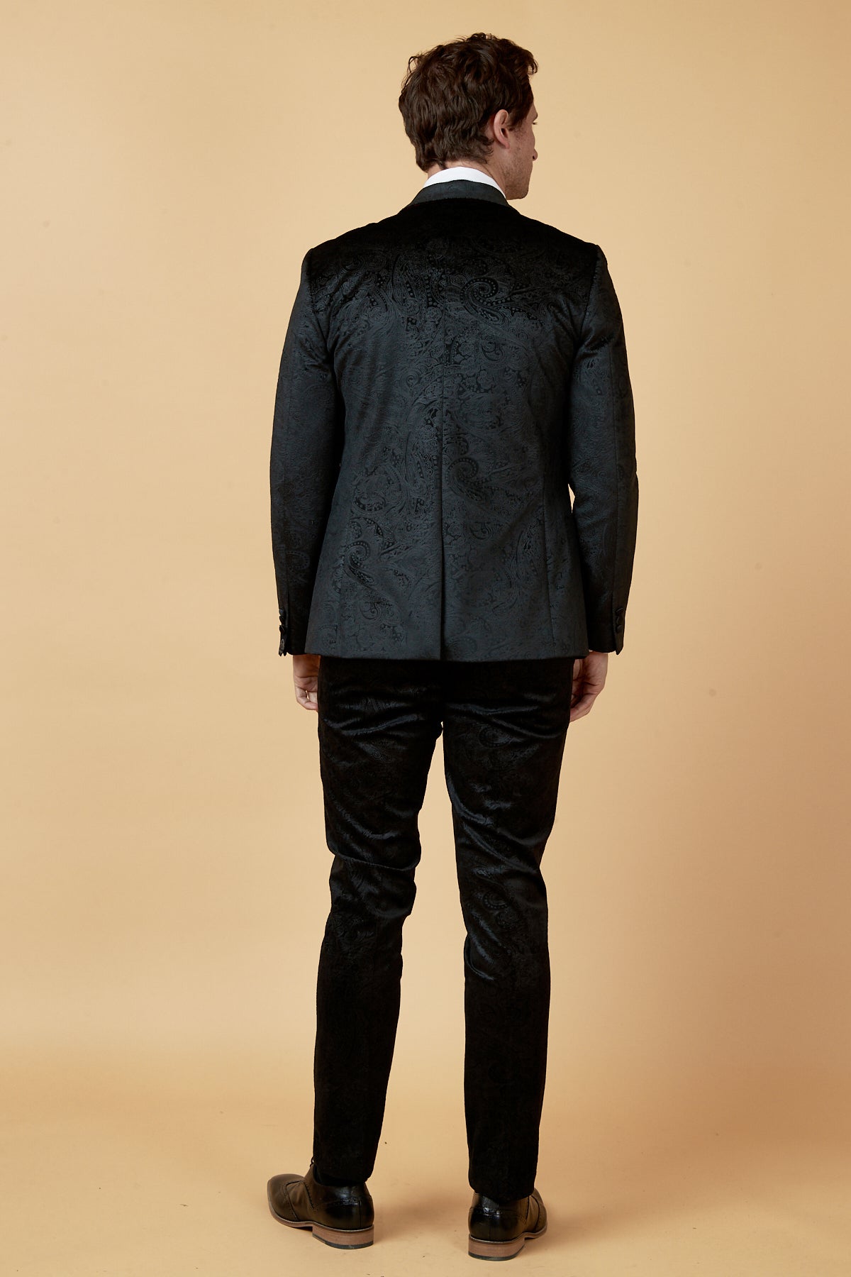 A man faces away from the camera, wearing the Marc Darcy SIMON Black Velvet Jacquard Three Piece Suit with a white shirt and dark shoes against a beige background—an ideal pick for elegant party wear.