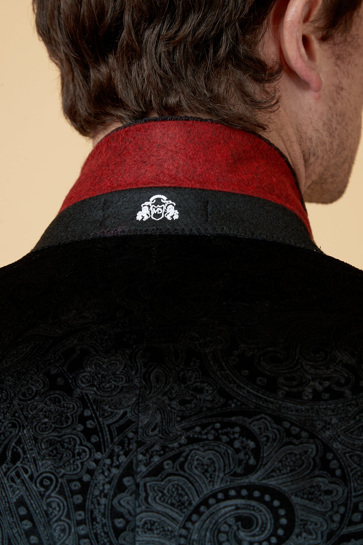 A close-up of a person’s upper back highlights the SIMON Black Velvet Jacquard Three Piece Suit by Marc Darcy, featuring ornate paisley patterns, a red collar, and a small white embroidered crest at the back.