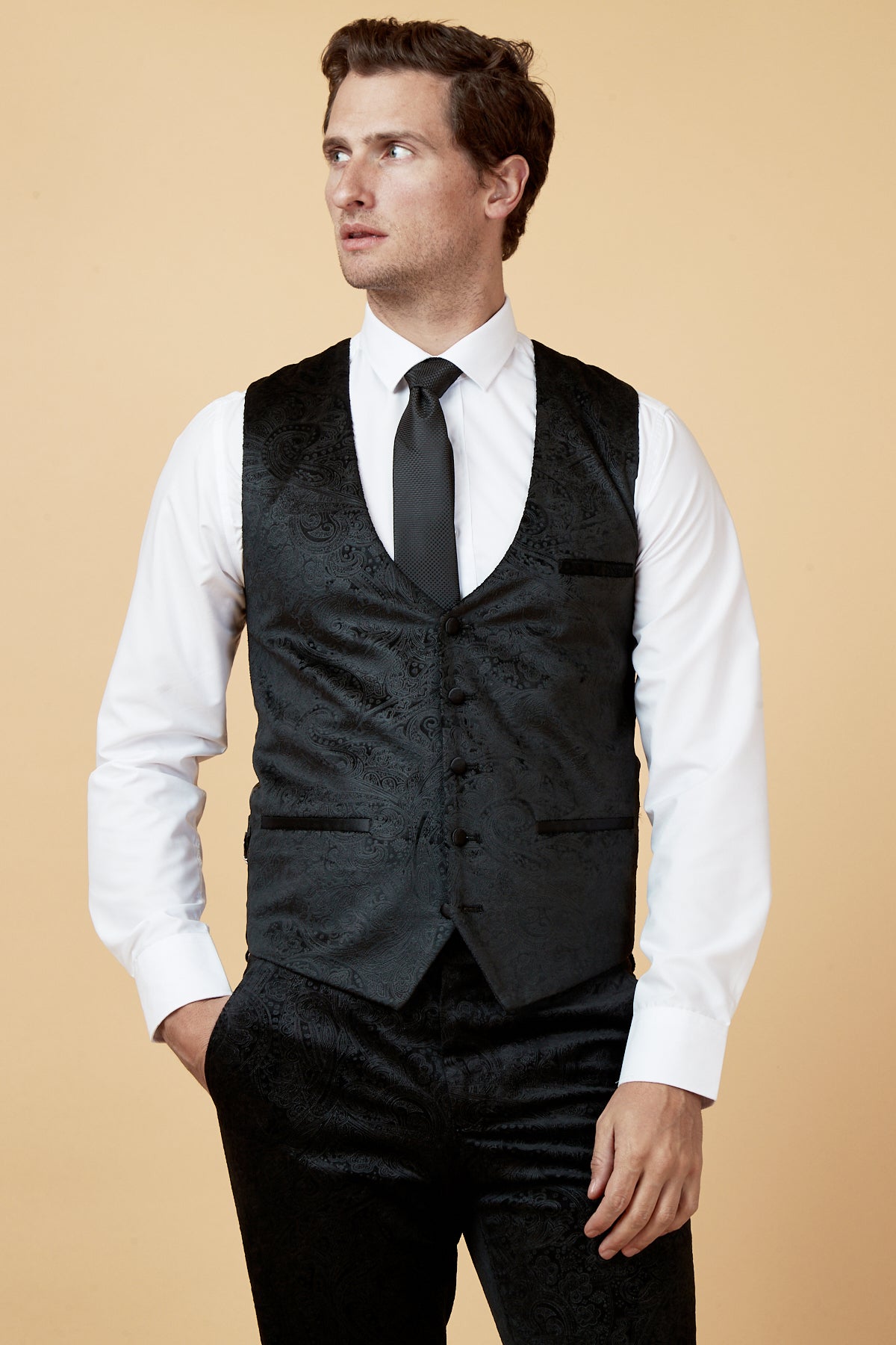 A man wears a marcdarcy SIMON Black Velvet Jacquard Waistcoat over a white dress shirt with matching black pants and tie, standing against a beige background, looking left with one hand in his pocket.