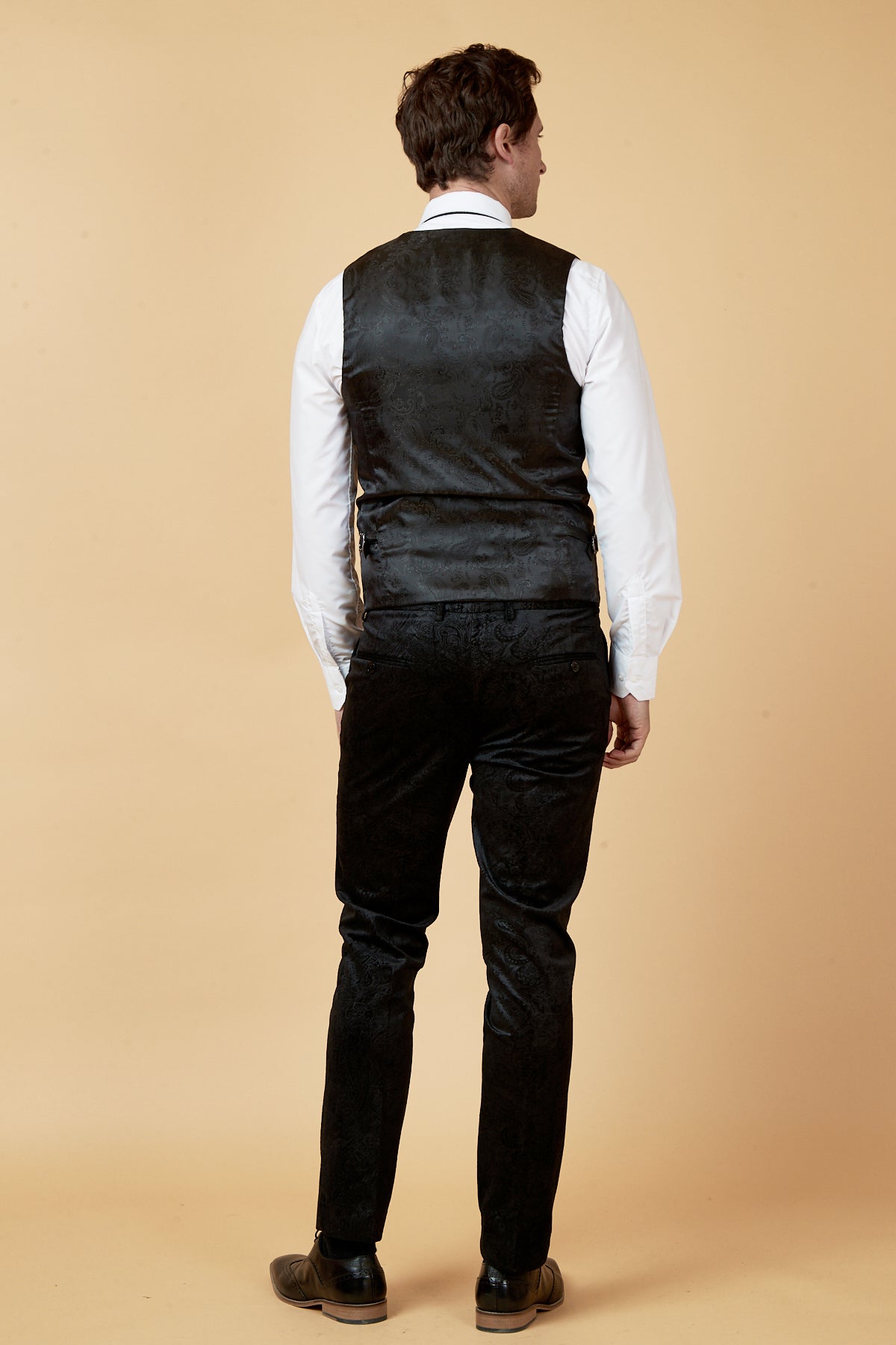 A man stands facing away from the camera, wearing a white dress shirt and black marcdarcy SIMON - Black Velvet Jacquard Waistcoat, paired with black dress pants and shoes against a plain beige background.