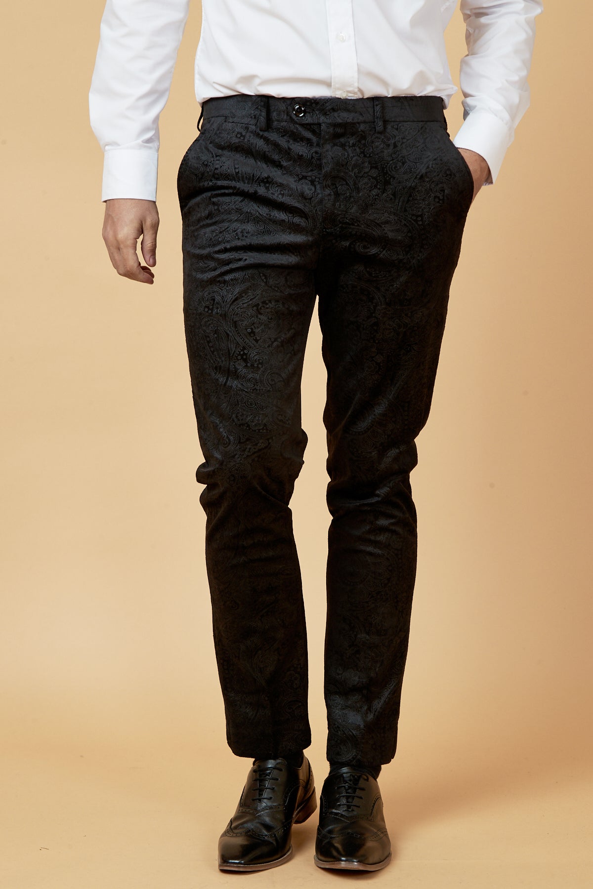 A man wears Marc Darcy's SIMON Black Velvet Jacquard Trousers, a white long-sleeve button-up shirt, and black dress shoes, posing with one hand in his pocket against a beige background—perfect for party dressing.