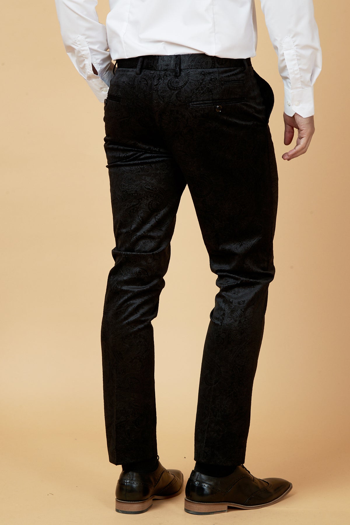 A man stands facing away, wearing a white dress shirt tucked into Marc Darcys SIMON Black Velvet Jacquard Trousers with an embossed paisley design and black dress shoes, set against a beige background.