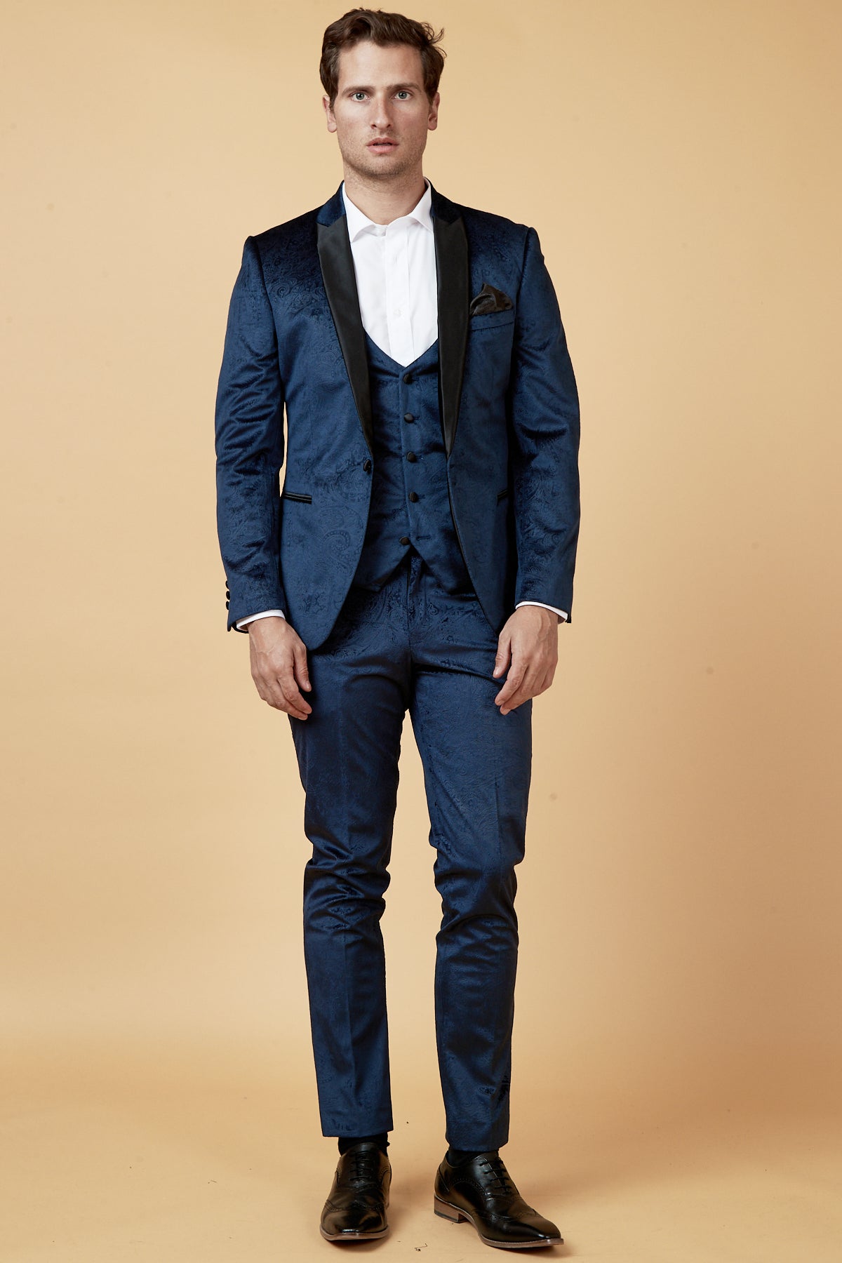 A man faces forward in a Marc Darcy SIMON Navy Velvet Jacquard Three Piece Suit, paired with a white dress shirt and black shoes, set against a plain beige background.