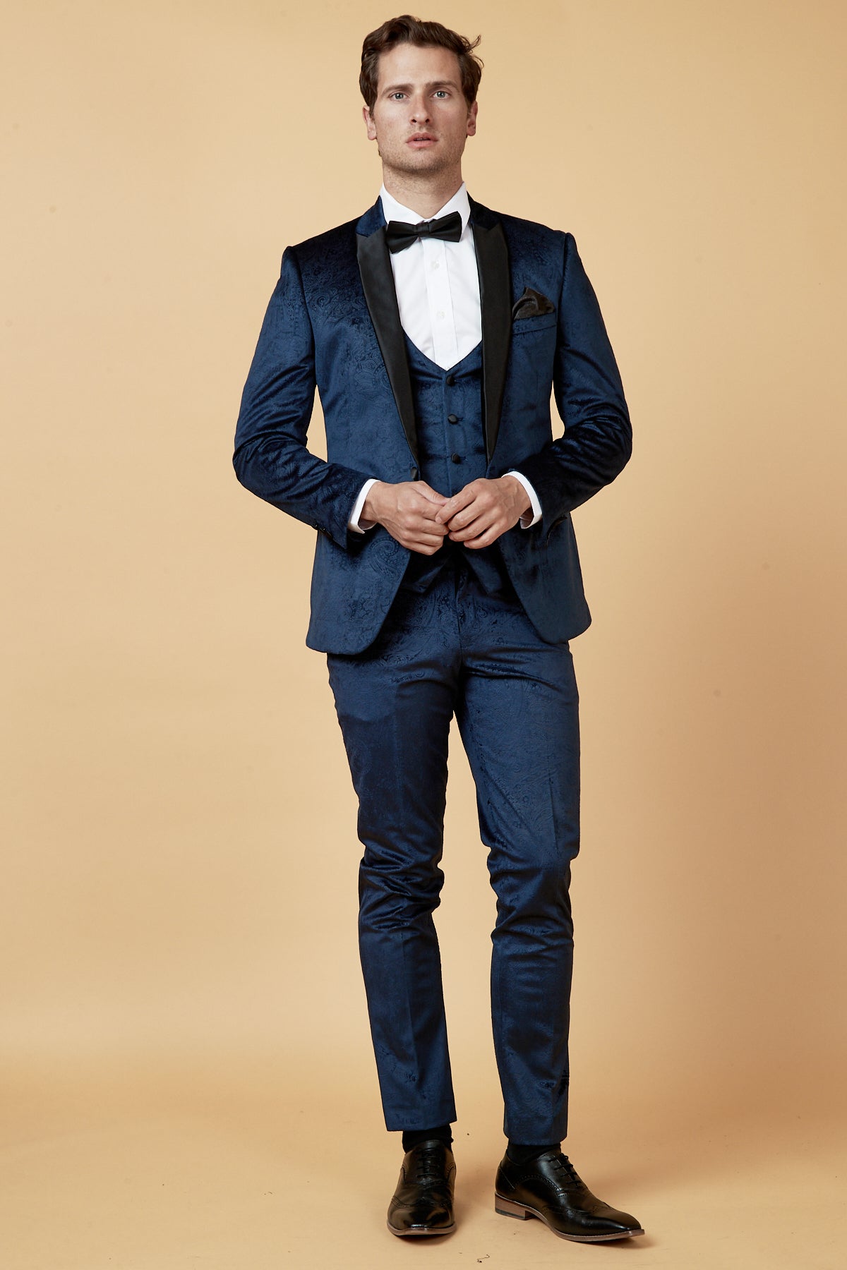 A man poses against a beige background in the Marc Darcy SIMON Navy Velvet Jacquard Three Piece Suit, styled with a white dress shirt, black bow tie and shoes, plus a matching blue vest and pocket square.