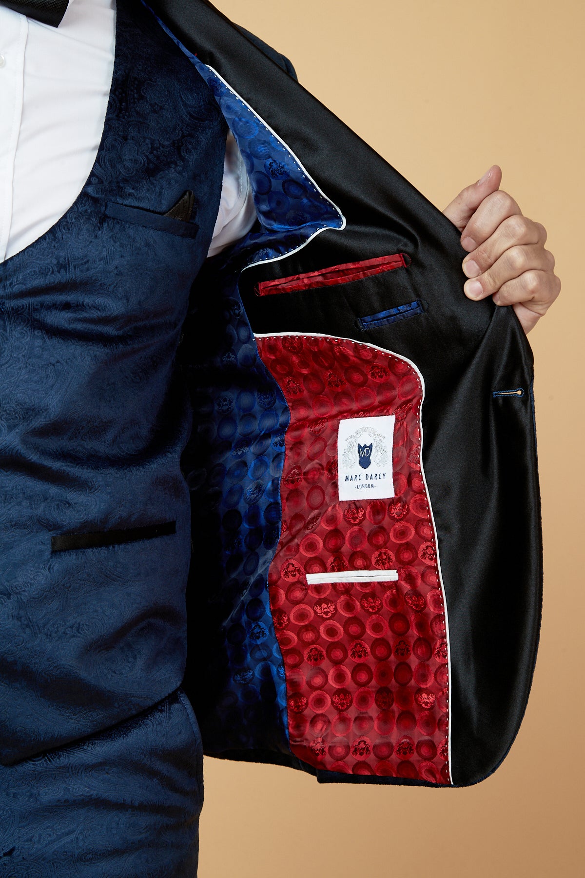 A person in the SIMON - Navy Velvet Jacquard Three Piece Suit by Marc Darcy holds open the jacket to show a patterned red and blue satin lining, multiple pockets, and a white Marc Darcy label.