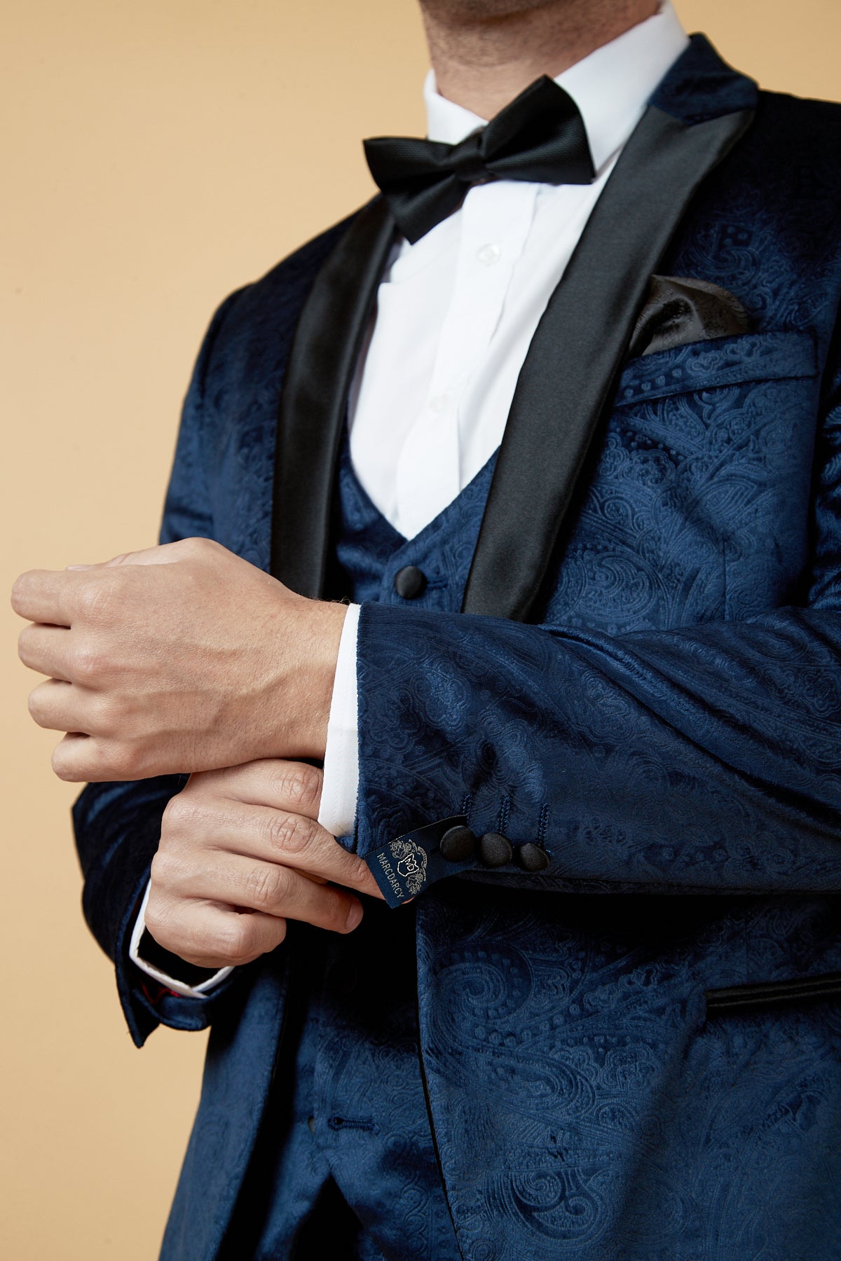 A man in the Marc Darcy SIMON Navy Velvet Jacquard Three Piece Suit with black lapels, white shirt, black bow tie, and pocket square adjusts his cufflink against a beige background.