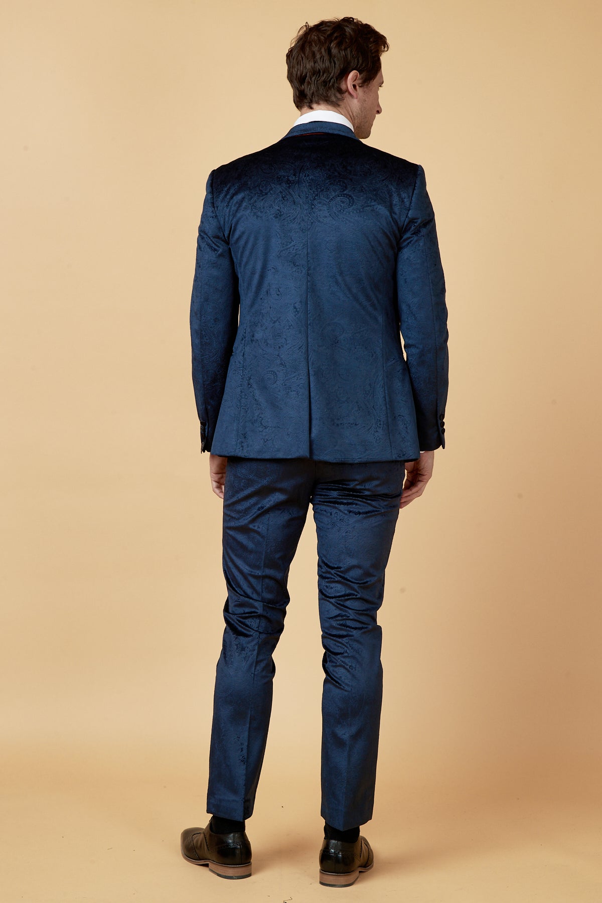 A man faces away from the camera, wearing the Marc Darcy SIMON Navy Velvet Jacquard Three Piece Suit with a white shirt and brown shoes, set against a plain beige background.