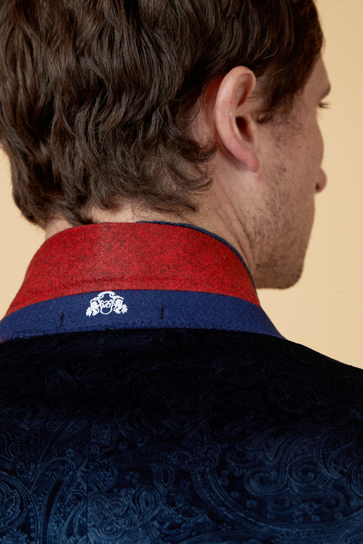 A close-up of a man's head and shoulders highlights the Marc Darcy SIMON Navy Velvet Jacquard Three Piece Suit, featuring a slim fit, red and blue collar, and white crest detail near the neckline against a beige background.
