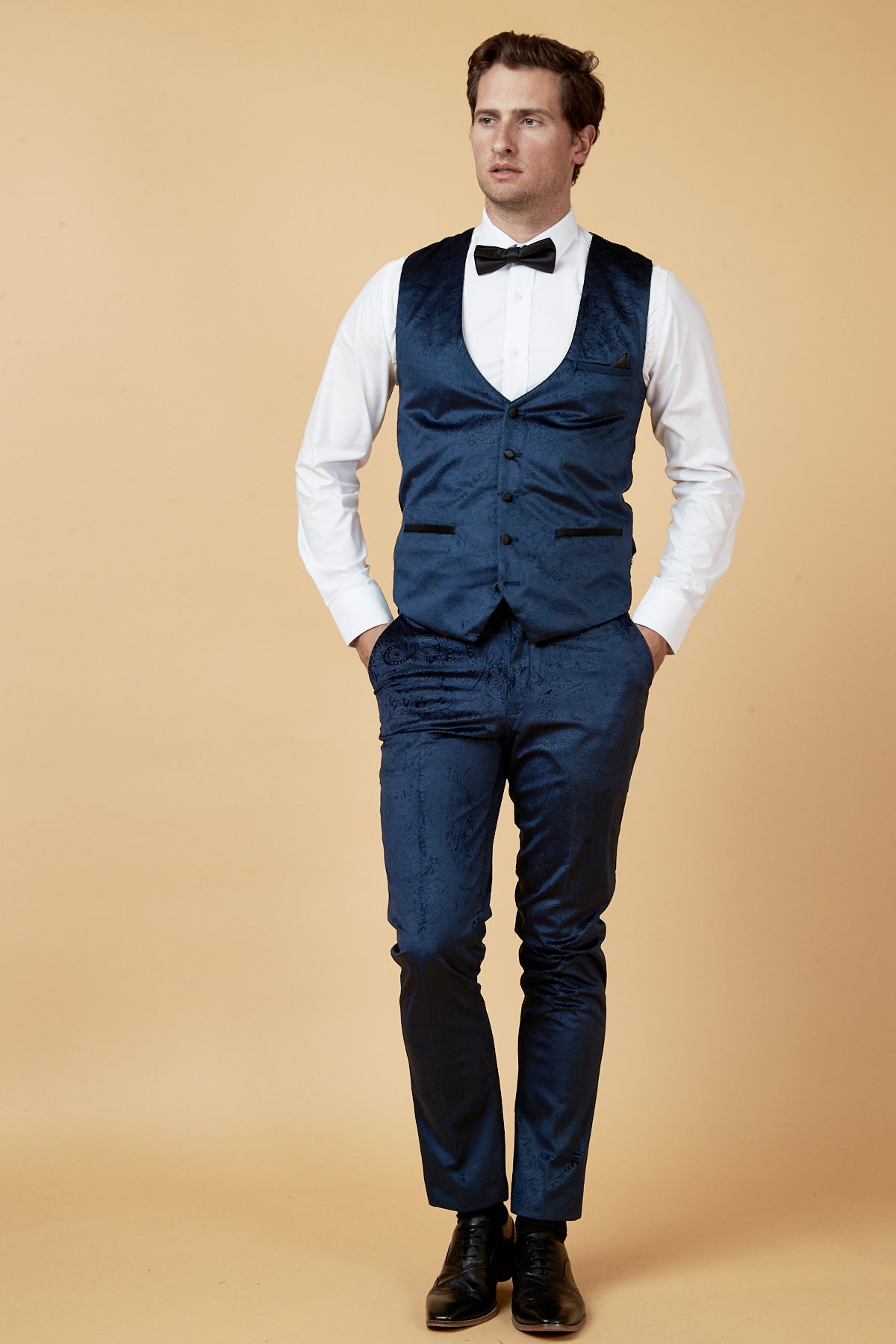 A man stands against a beige background, wearing the marcdarcy SIMON navy velvet jacquard waistcoat with matching pants, a white dress shirt, black bow tie, and black dress shoes, his hands in his pockets.