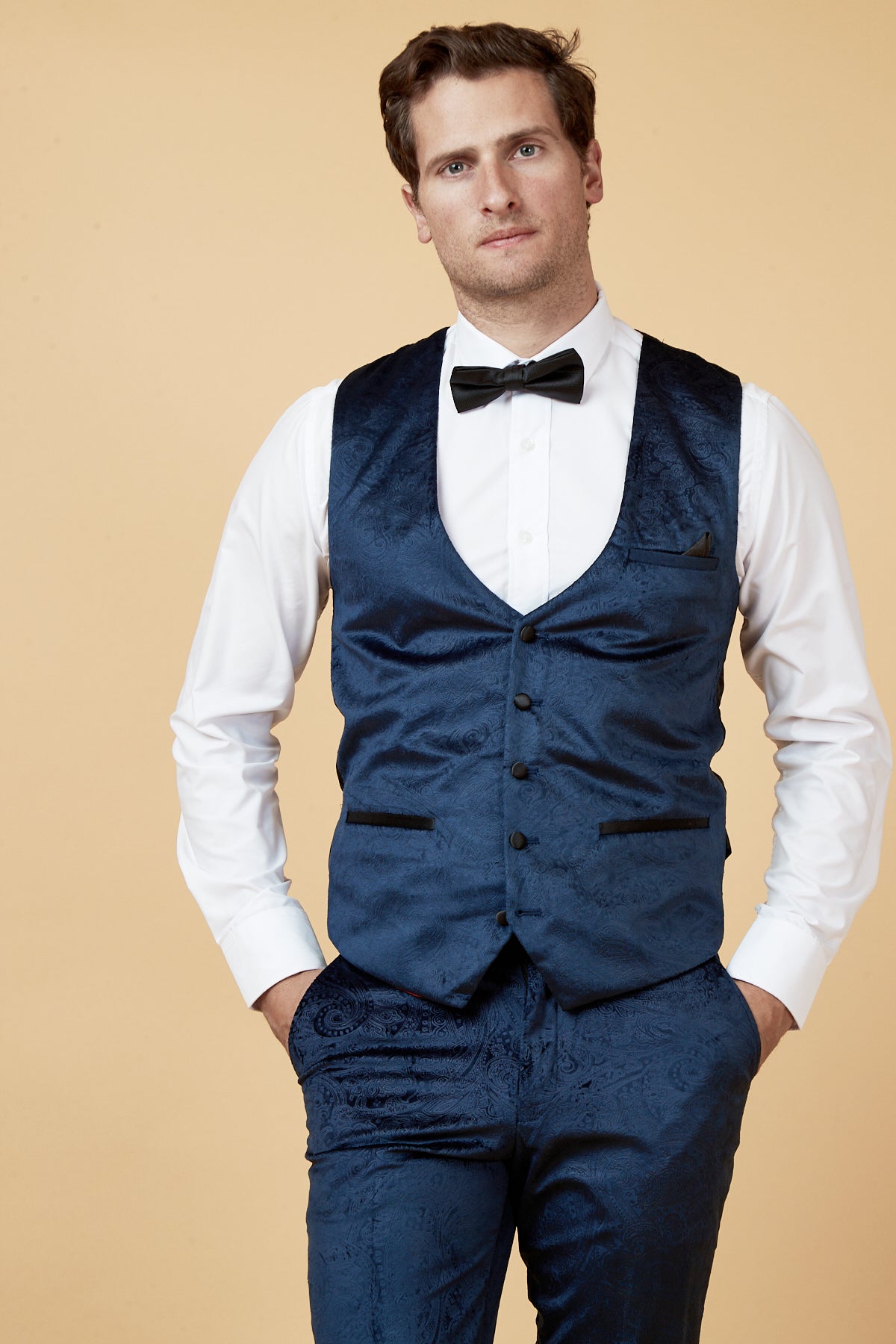 A man wears the Marc Darcy SIMON Navy Velvet Jacquard Three Piece Suit, featuring a patterned waistcoat over a white dress shirt and black bowtie, with matching blue trousers. His hands are in his pockets.