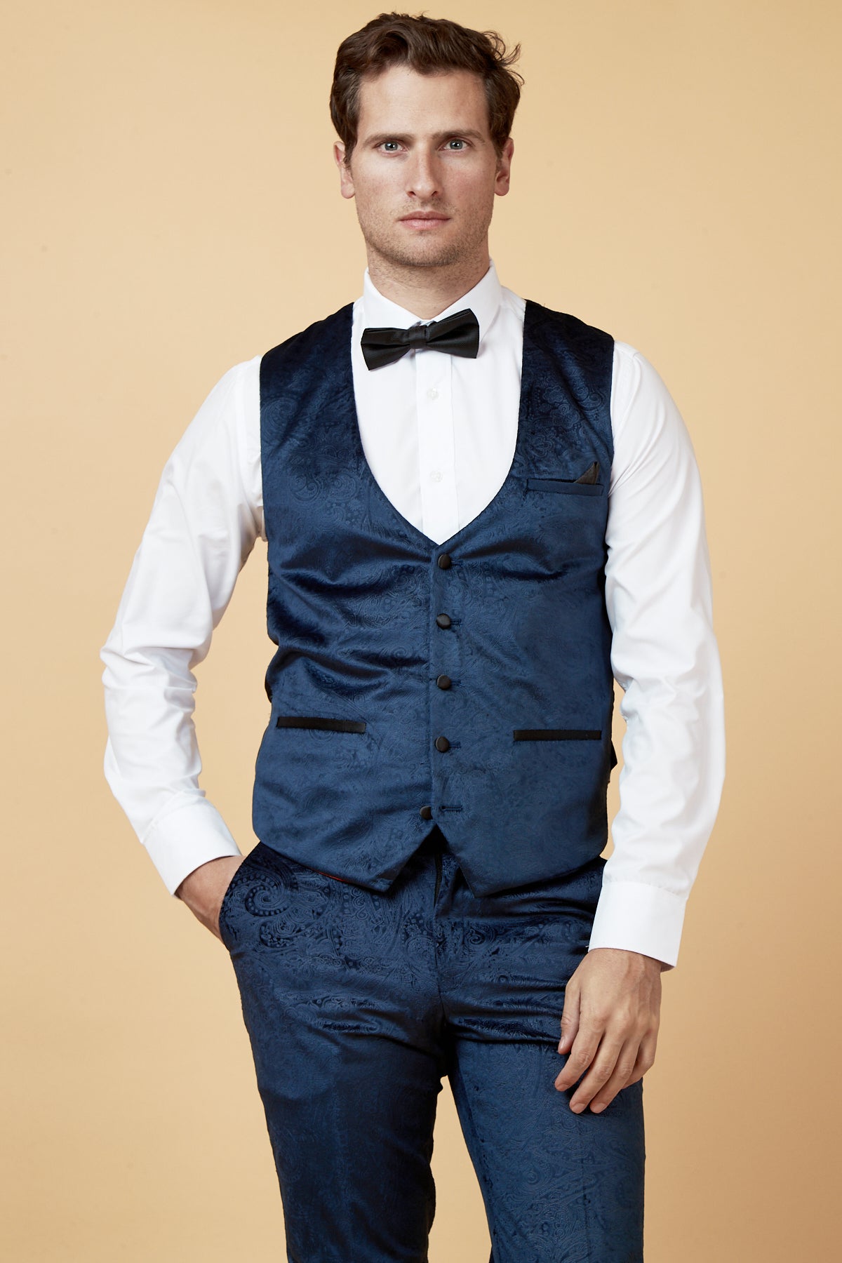 A man stands against a beige background wearing a white dress shirt, black bow tie, marcdarcy’s SIMON Navy Velvet Jacquard Waistcoat, and matching blue paisley brocade pants, one hand in his pocket as he looks confidently at the camera.