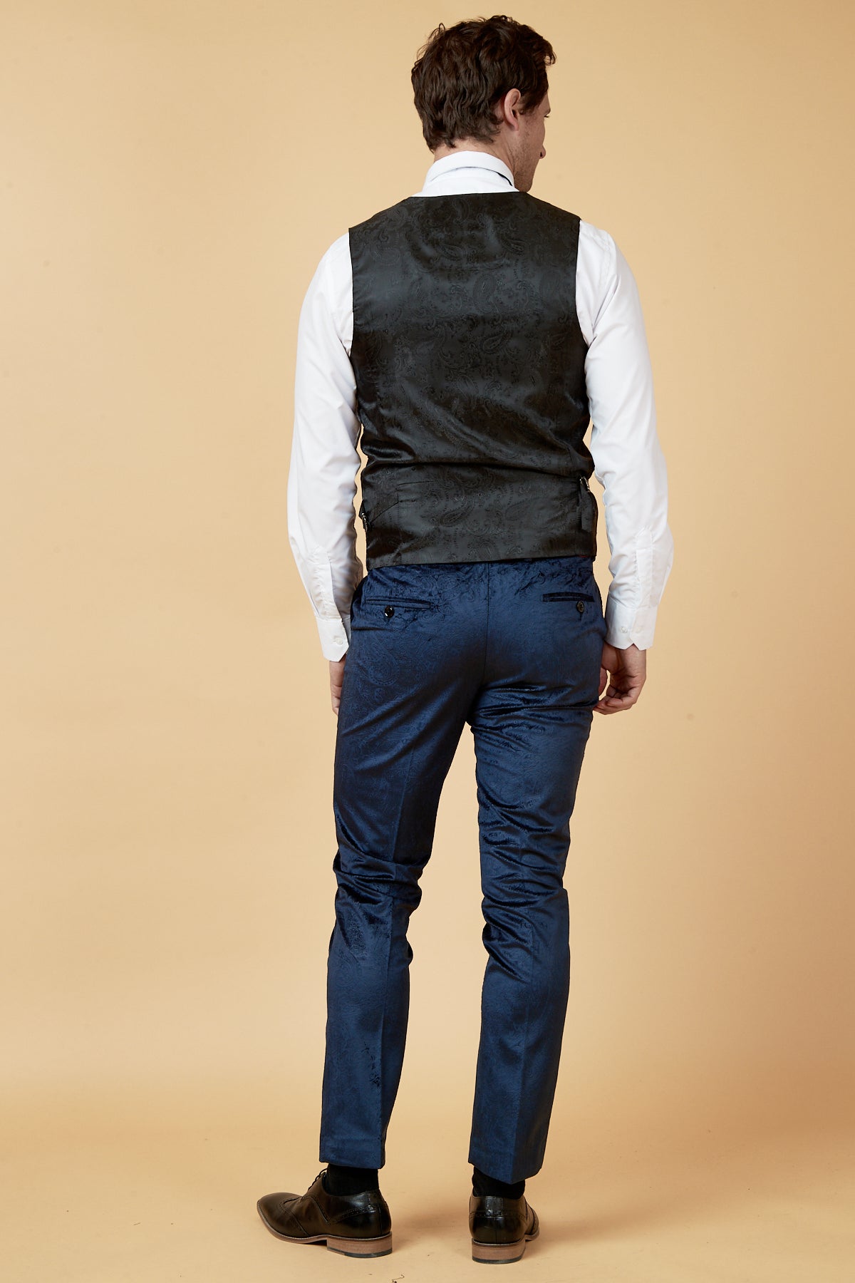 A man stands facing away from the camera, wearing a white dress shirt, marcdarcy SIMON Navy Velvet Jacquard Waistcoat, blue dress pants, and dark shoes against a plain beige background.