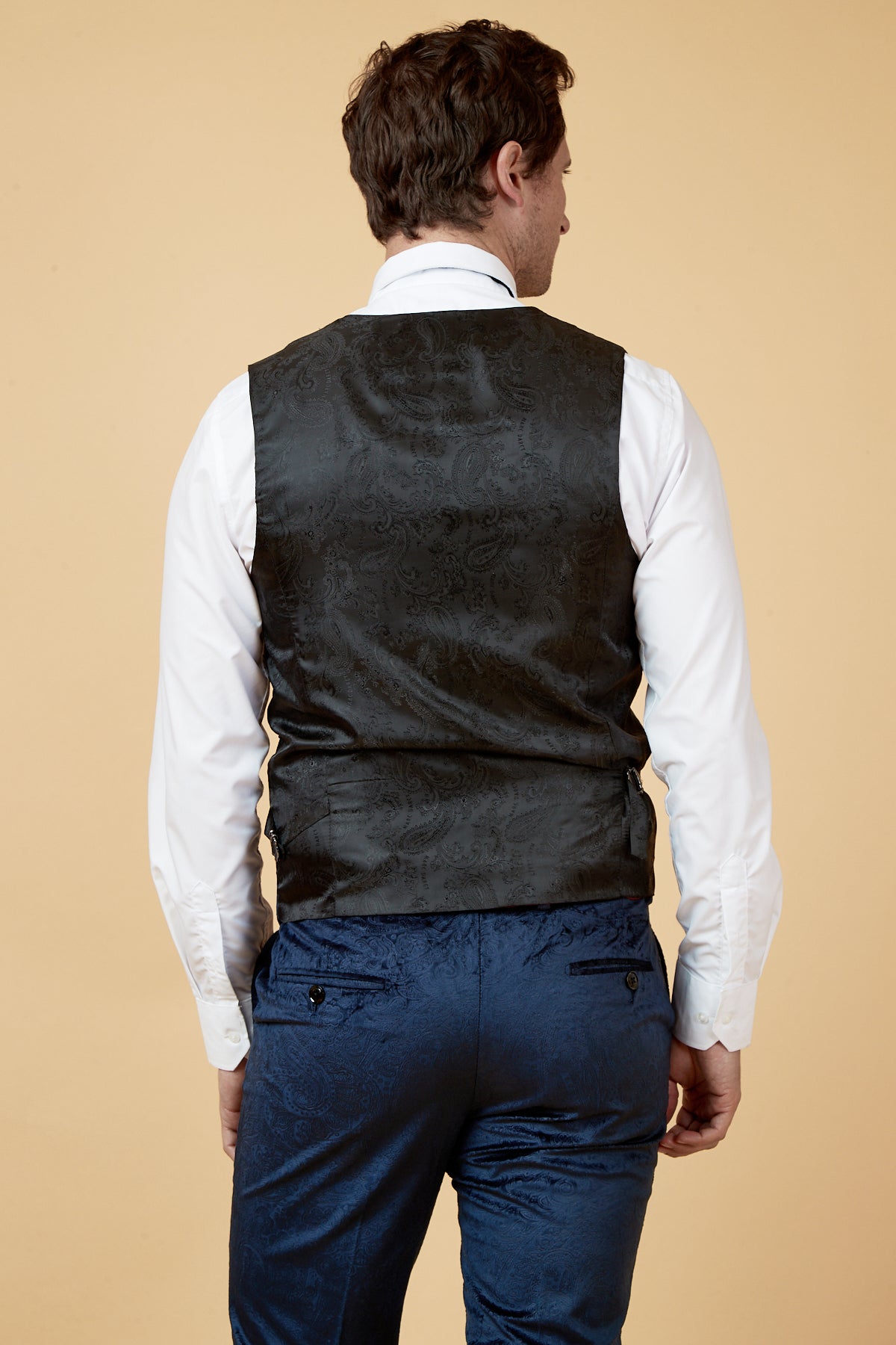 A man seen from the back wears Marc Darcy's SIMON Navy Velvet Jacquard Three Piece Suit, featuring a slim fit navy paisley vest over a white shirt and matching blue paisley pants, set against a beige background.