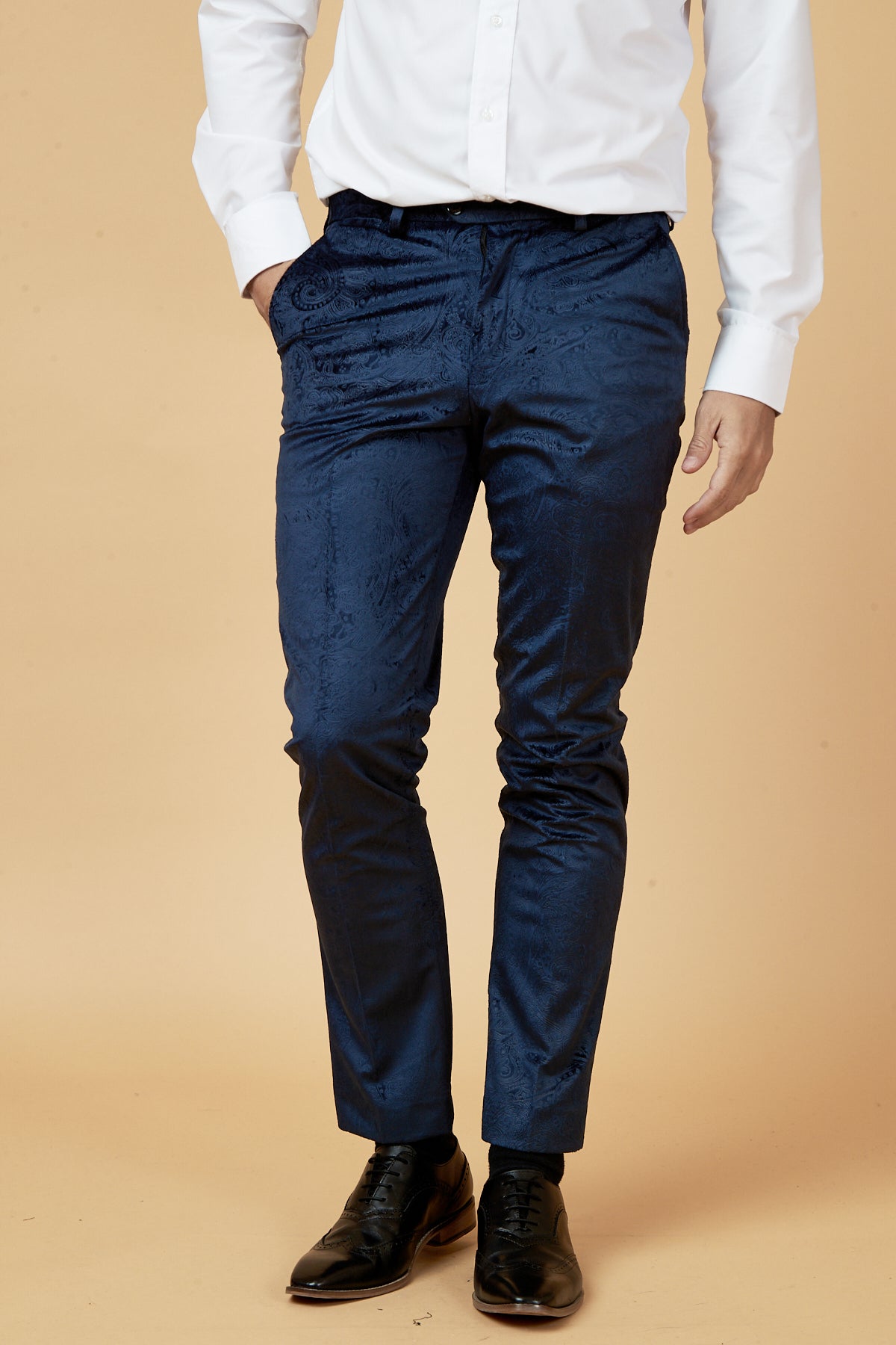 A man models the Marc Darcy SIMON Navy Velvet Jacquard Three Piece Suit with a white dress shirt and black shoes against a beige background, one hand in his pocket. His head is cropped out of the image.