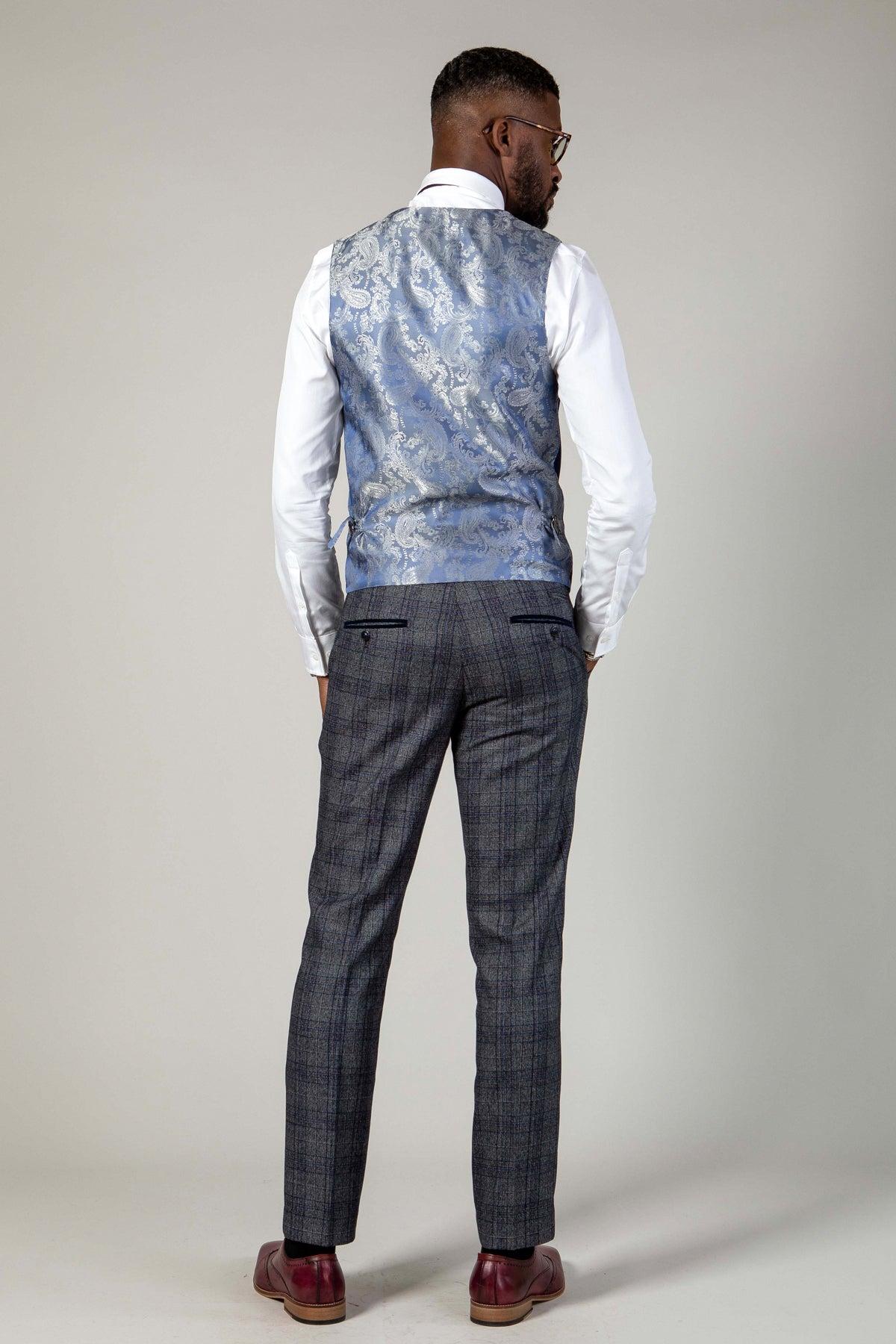 A man stands with his back to the camera, wearing the marcdarcy KELVIN navy double-breasted waistcoat, a white shirt, gray plaid trousers, and burgundy shoes against a plain light-colored background.