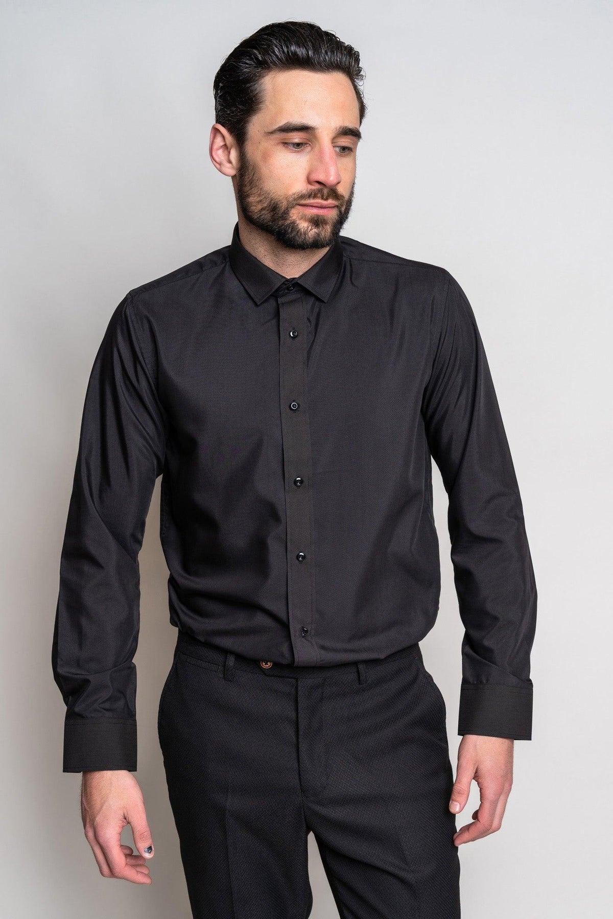 A man with dark hair and a beard wears the Marc Darcy CARTER Black Long Sleeve Shirt and black pants, standing against a plain light background. He looks slightly down and to his left with a neutral expression.