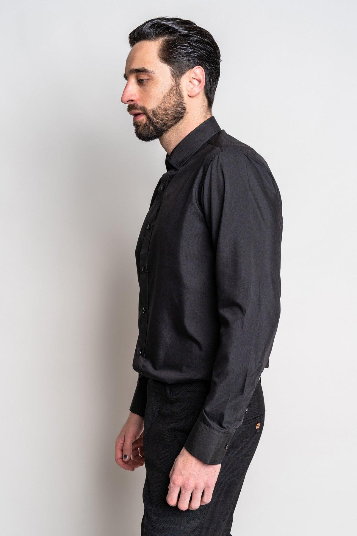 A man with dark hair and a beard stands in profile against a plain light background, wearing the Marc Darcy CARTER black long sleeve shirt with a spread collar and dark pants.
