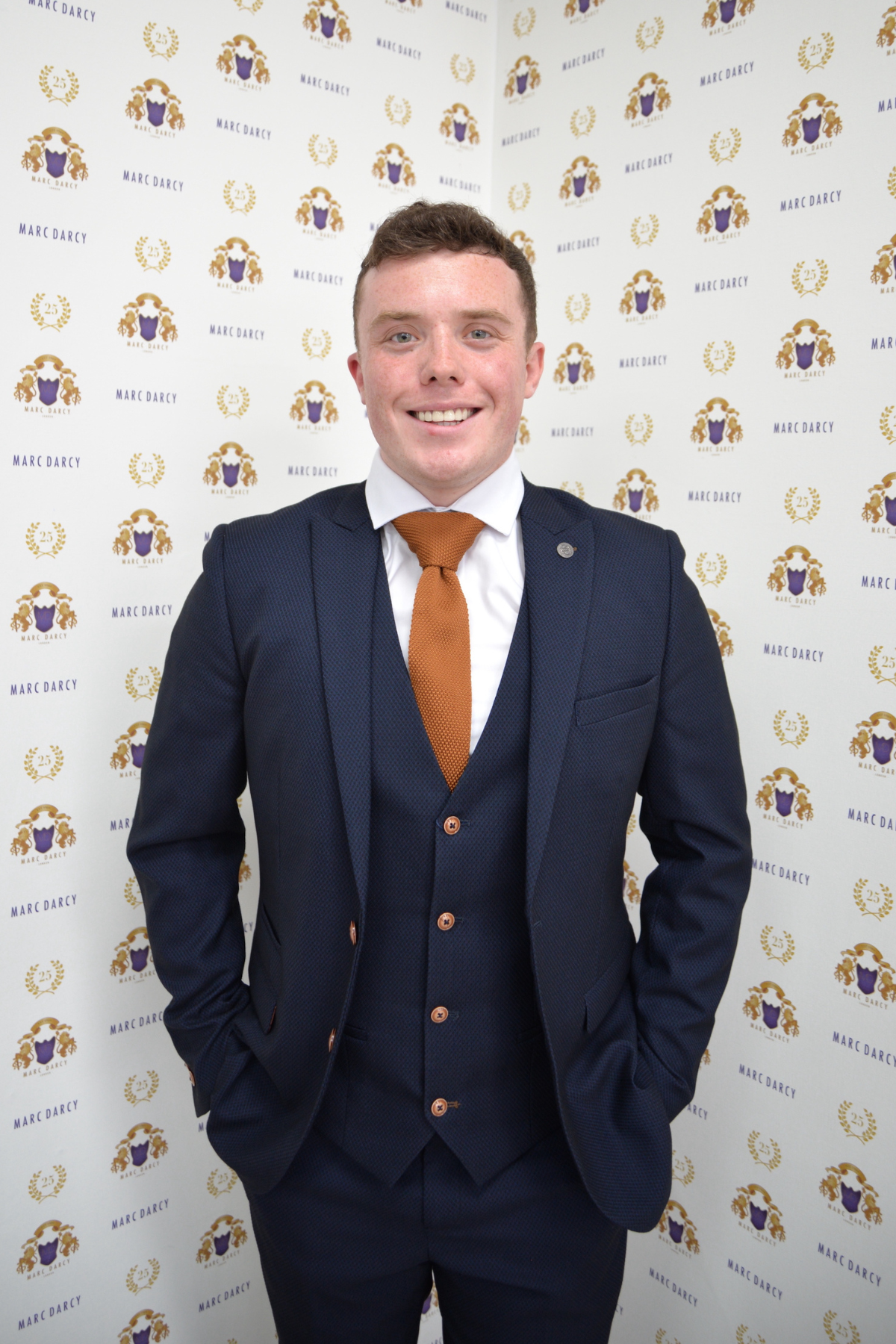 Emmerdale's Bradley Johnson (Vinny Dingle) smiles in front of a patterned backdrop, wearing the sharp slim-fit Max Navy Suit by marcdarcy.