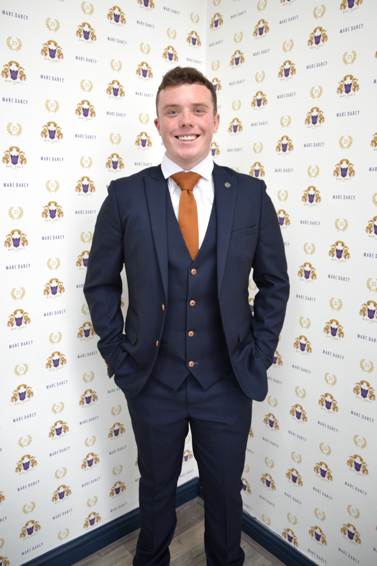 Emmerdale’s Bradley Johnson (Vinny Dingle) wears the slim fit Max navy suit by marcdarcy, paired with a white shirt and orange tie, standing against a wall featuring the repeated “marcdarcy” and lion crest logo.