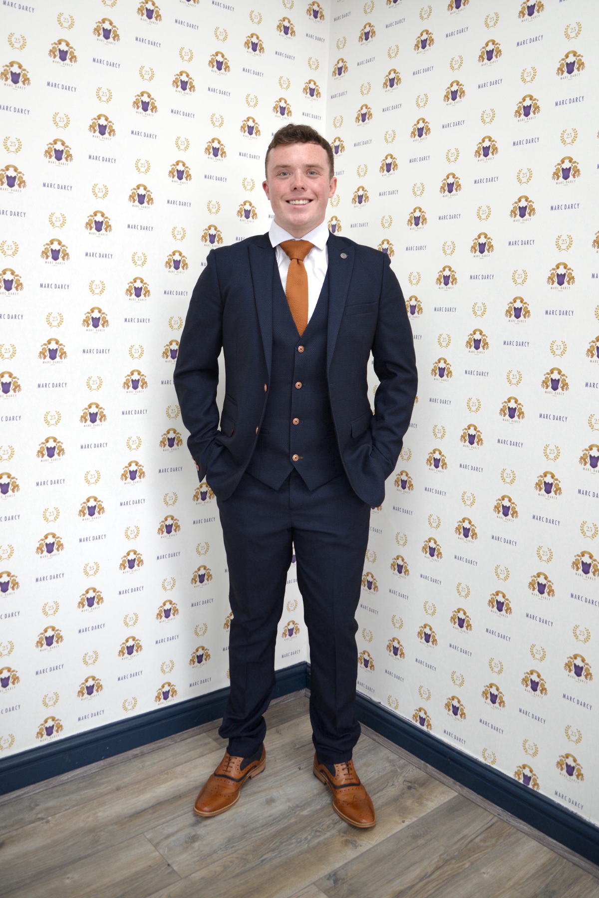 Emmerdale’s Bradley Johnson (Vinny Dingle) wears the marcdarcy Max Navy Slim Fit Suit with a white shirt and brown shoes, posing with hands in pockets before a logo wall on a wooden floor.