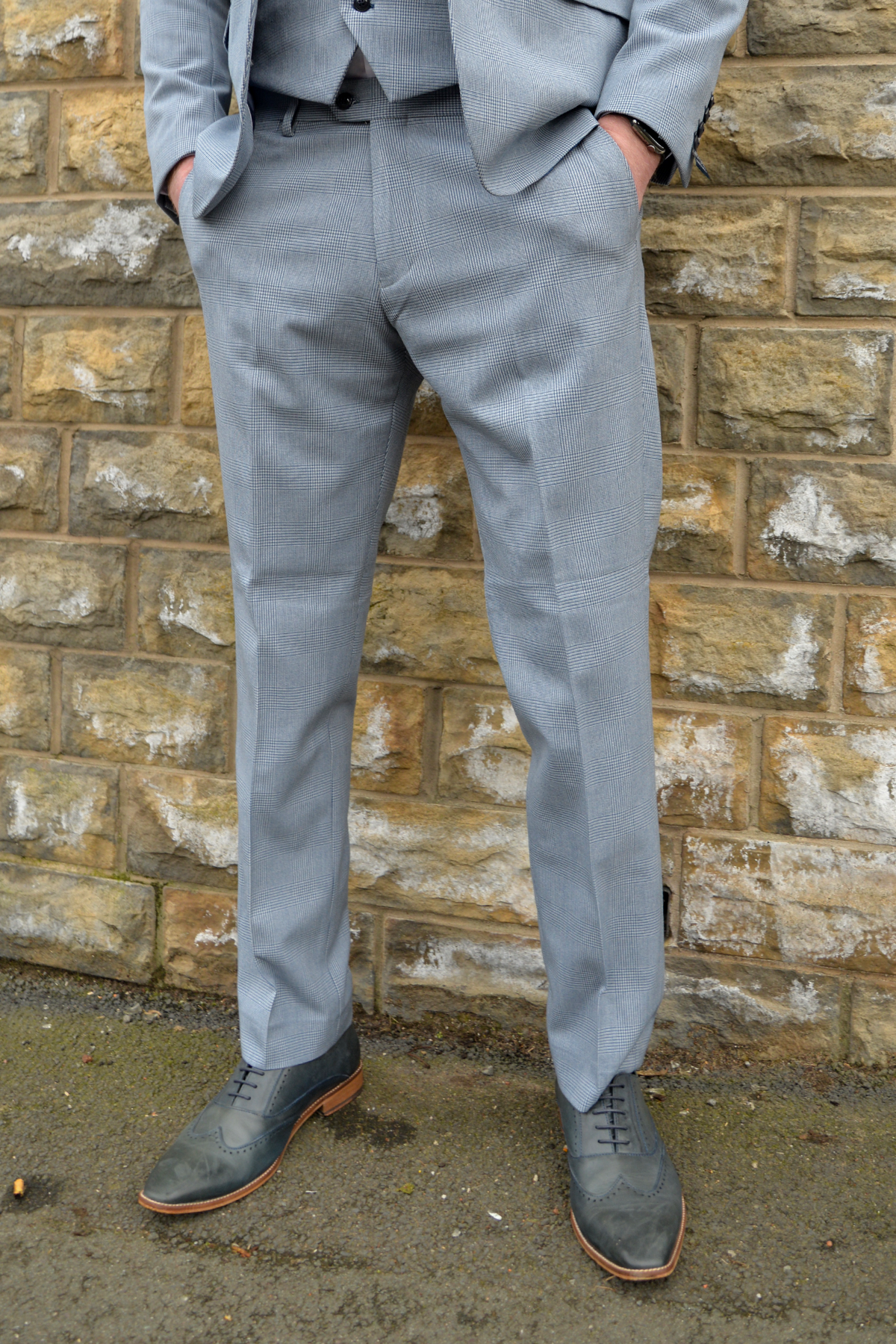 A man in The Everton Collection | James Garner In The BROMLEY Sky Blue Suit by Marc Darcy stands with his hand in his pocket against a brick wall.