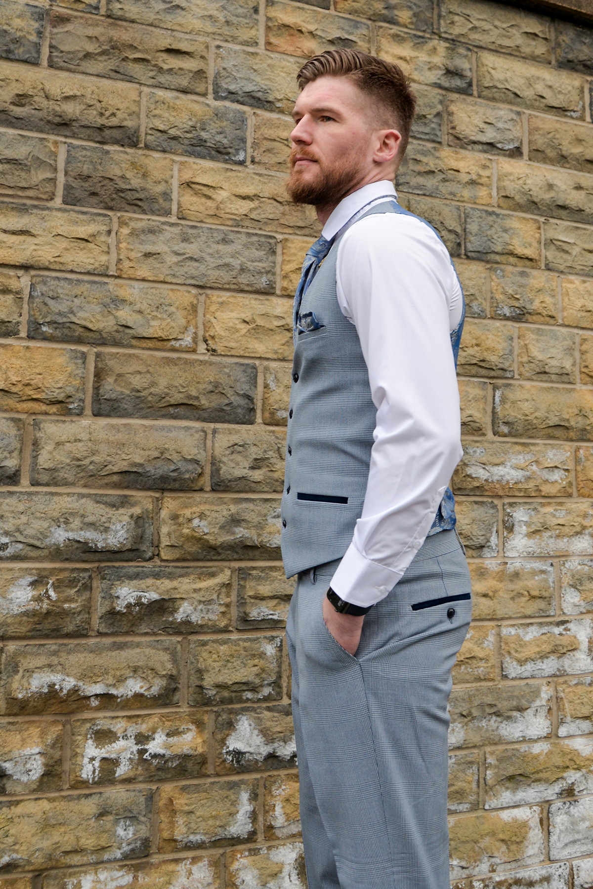 A man with short hair and a beard wears the Marc Darcy "James Garner In The BROMLEY Sky Blue Suit" from The Everton Collection, standing sideways against a stone brick wall and looking slightly away from the camera.