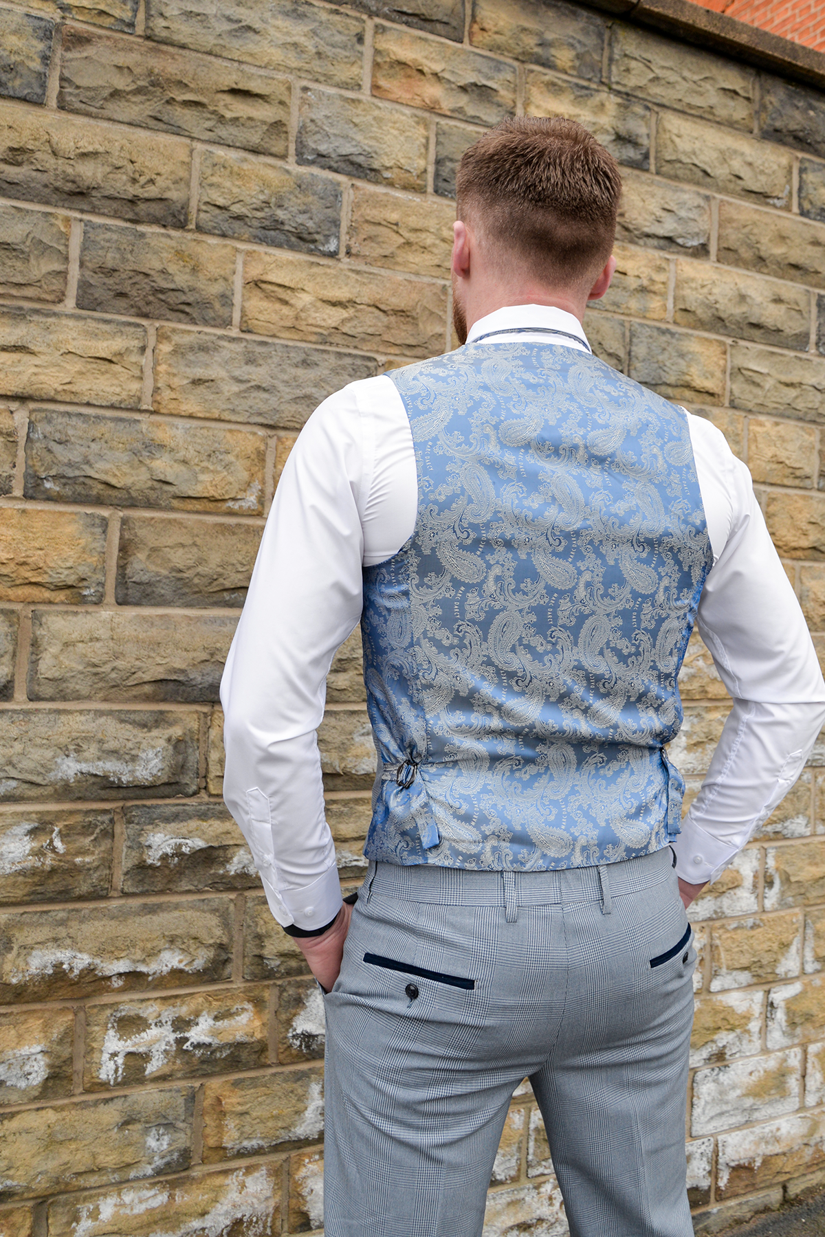 A man with short brown hair faces a brick wall, wearing The Everton Collection | James Garner In The BROMLEY Sky Blue Suit by Marc Darcy; his hands rest by his sides.