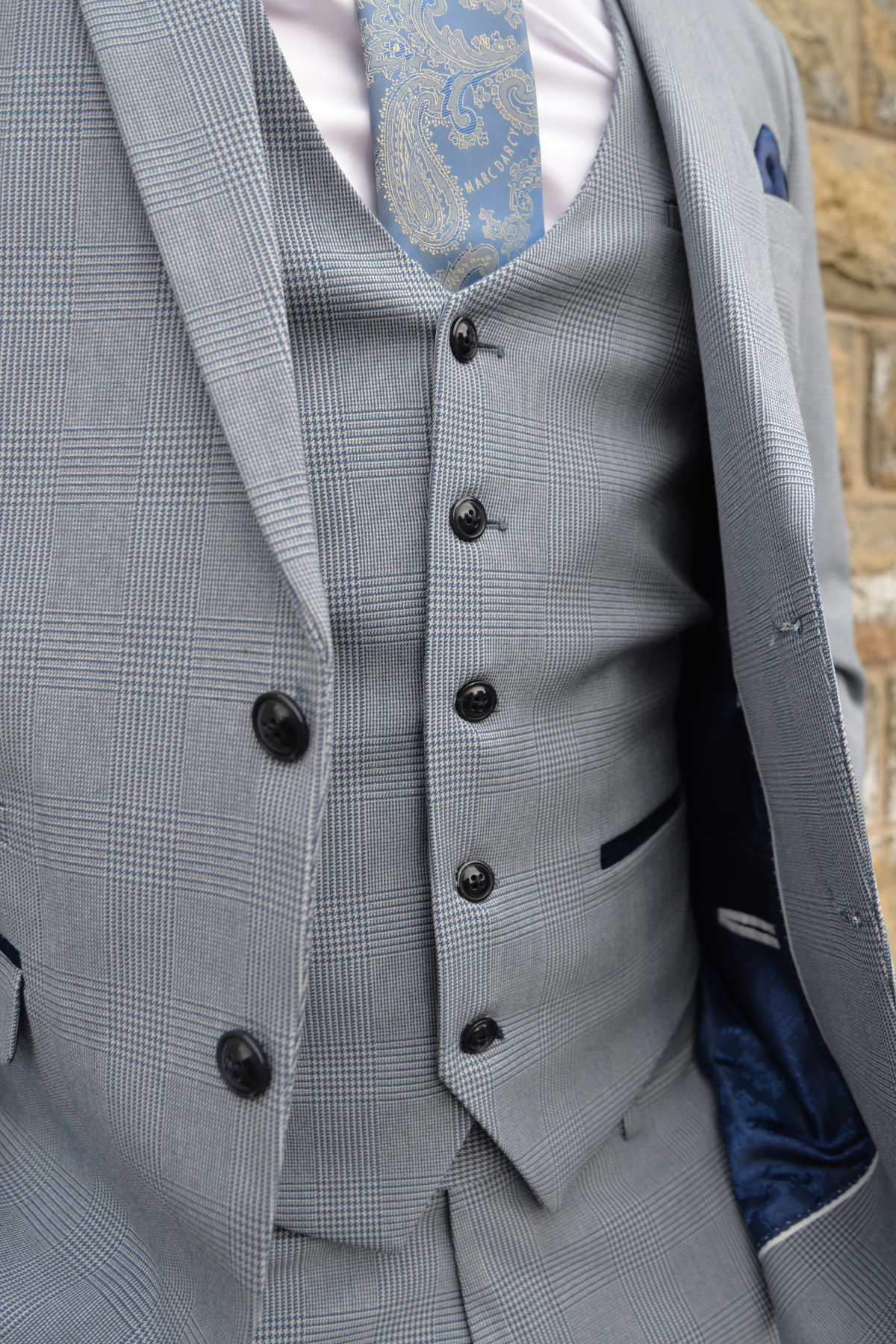 A close-up of a person in Marc Darcy’s The Everton Collection | James Garner In The BROMLEY Sky Blue Suit, standing by a stone wall.