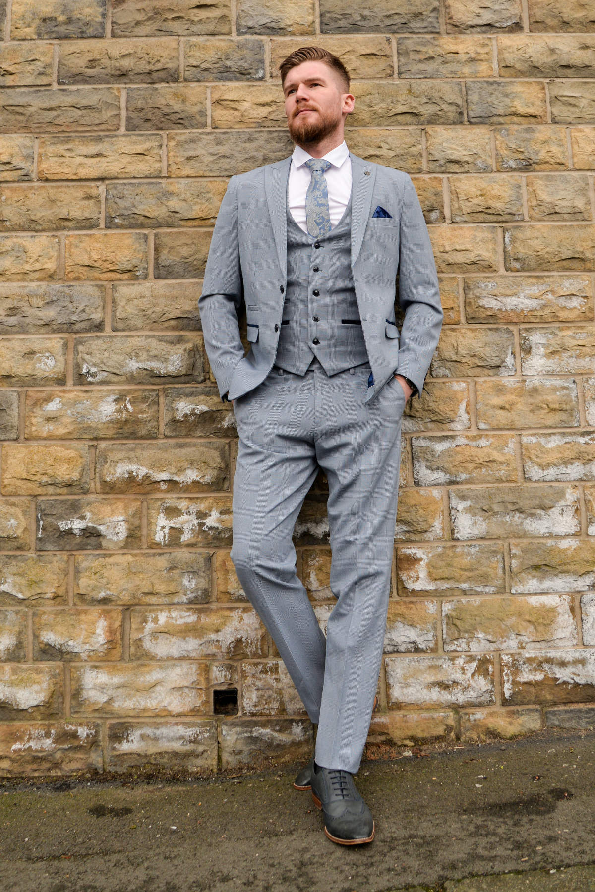 A man wearing The Everton Collection James Garner In The BROMLEY Sky Blue Suit by Marc Darcy stands against a stone wall with his hands in his pockets, looking to the side.