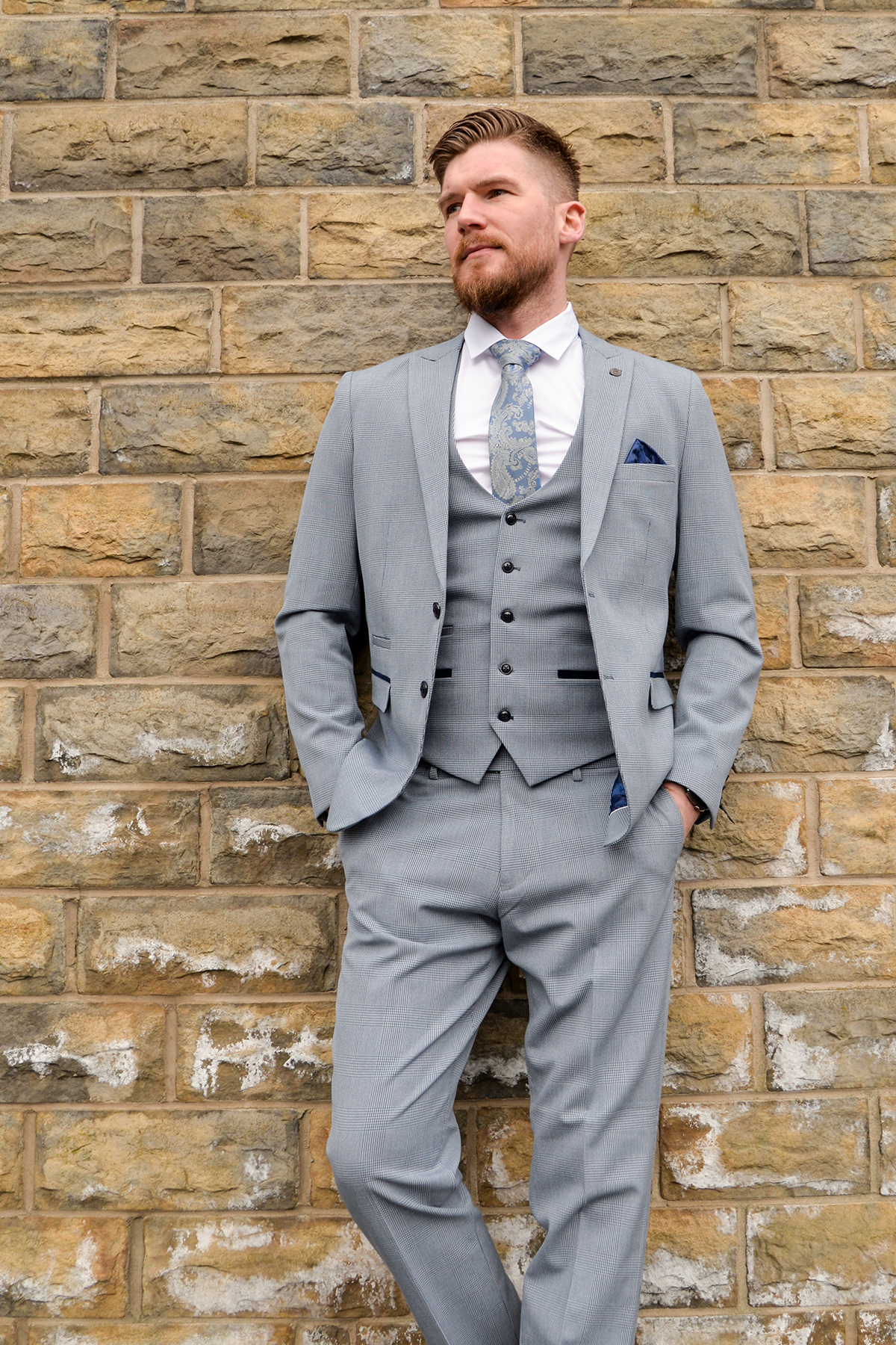 A man with short hair and a beard, wearing Marc Darcys The Everton Collection | James Garner In The BROMLEY Sky Blue Suit, stands against a stone brick wall with his hands in his pockets, gazing to the side.