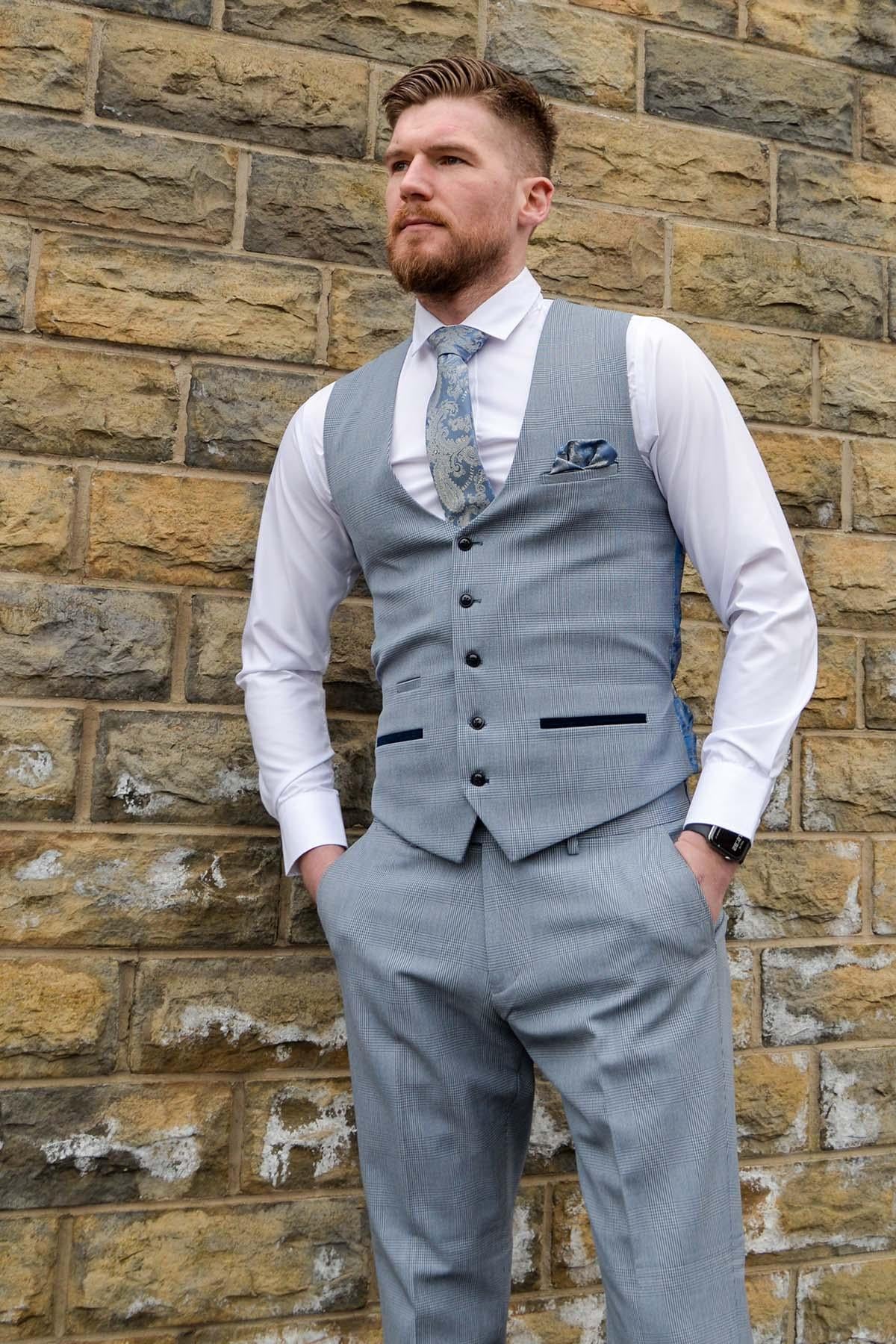 A bearded man in The Everton Collection | James Garner In The BROMLEY Sky Blue Suit by Marc Darcy stands against a stone wall with hands in pockets, wearing a white shirt and patterned tie, looking off to the side.