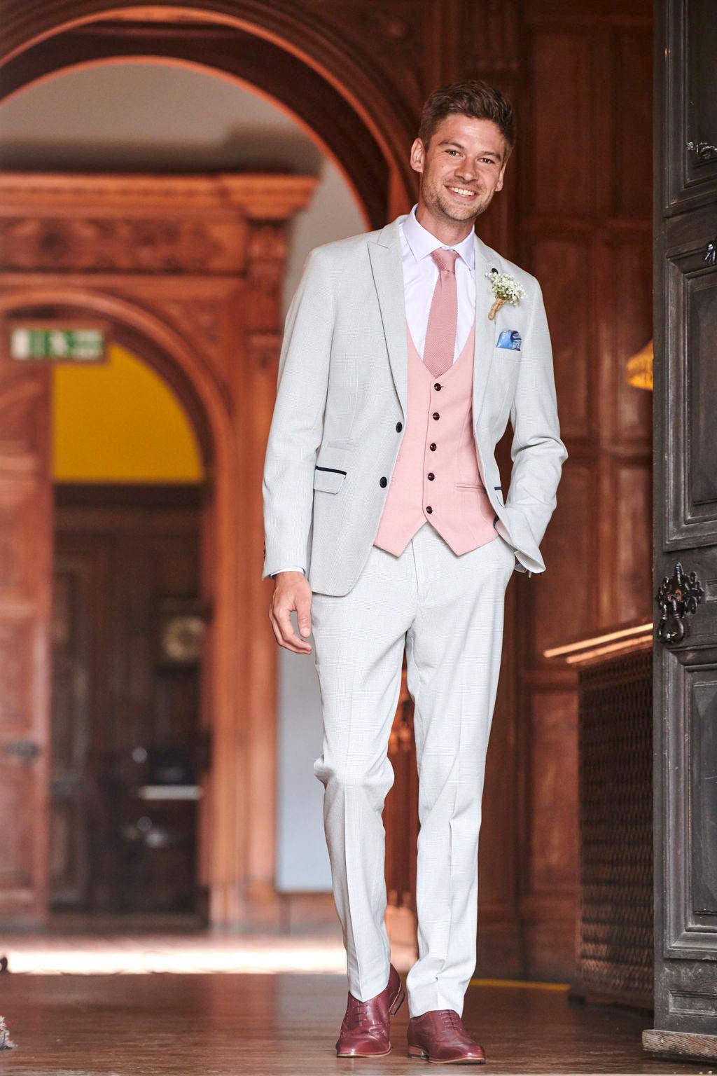 A smiling man in the Marc Darcy BROMLEY Stone Check Suit with Kelvin Pink Waistcoat walks indoors through an ornate wooden doorway, wearing a white shirt, brown shoes, and a boutonnière.