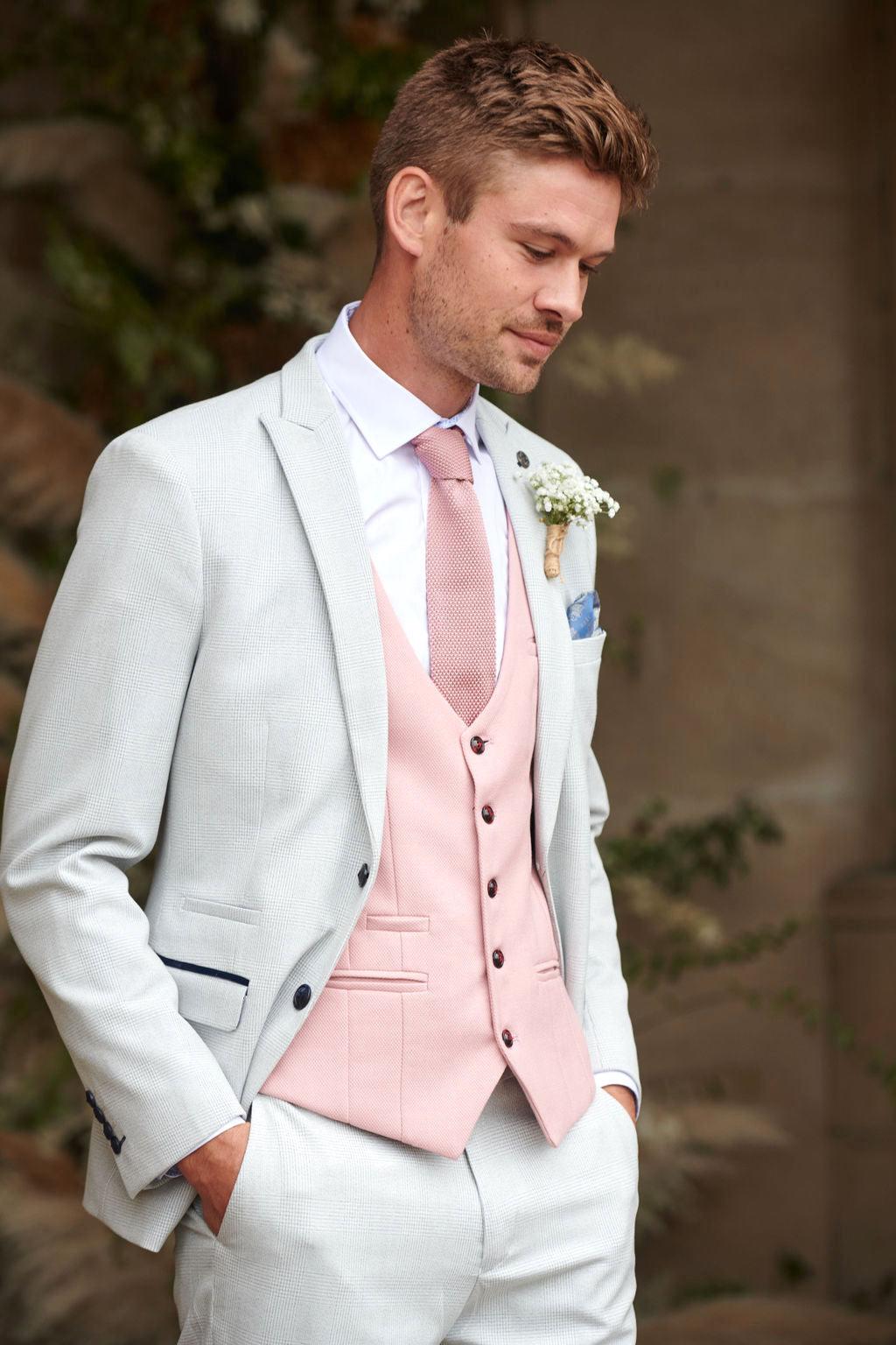 A man wears the Marc Darcy BROMLEY Stone Check Suit with a Kelvin Pink Waistcoat, matching tie, and white shirt. Hands in pockets, he looks down as a boutonnière decorates his lapel; blurred greenery forms the backdrop.