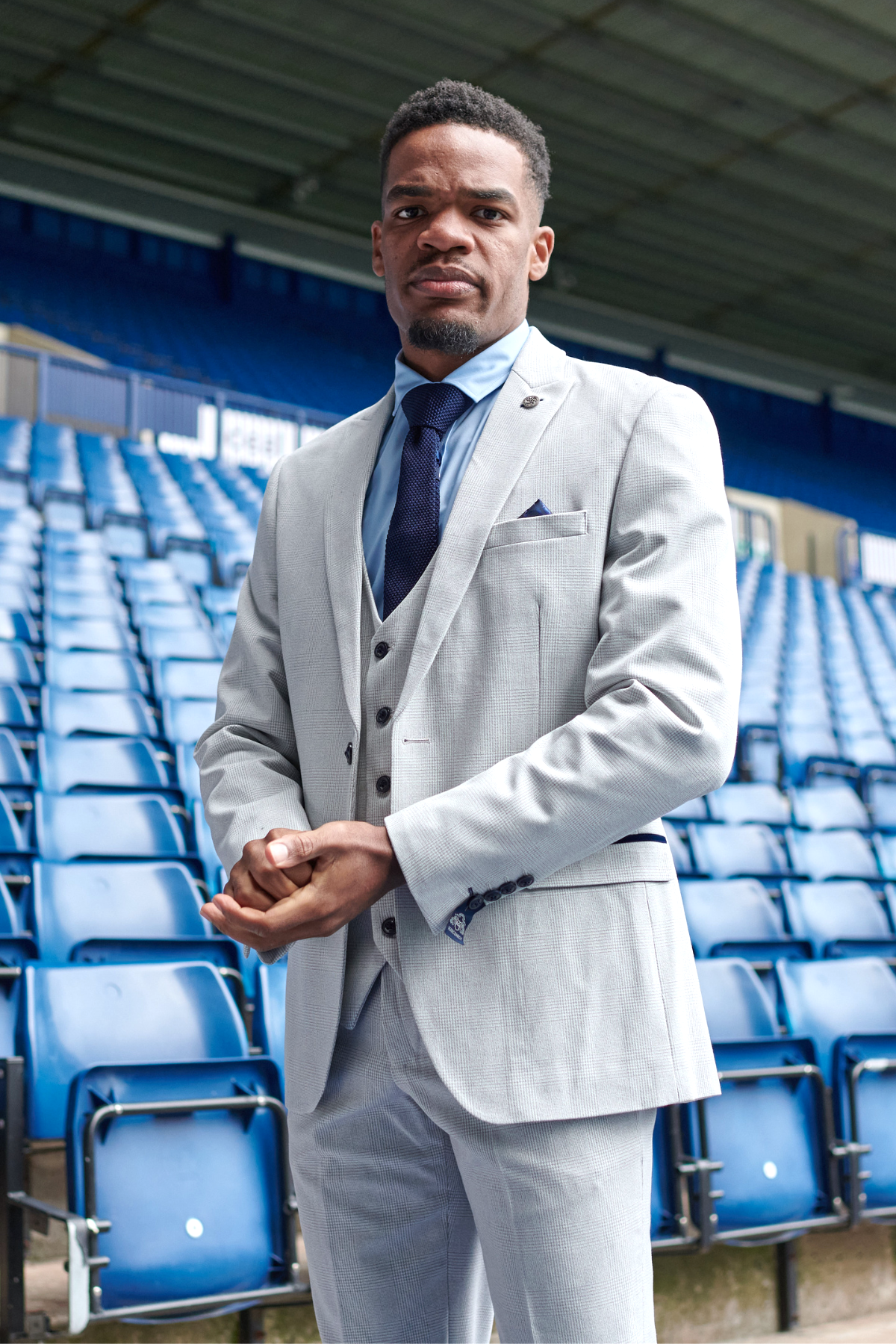 A man wears the Marc Darcy WBA Collection - Bromley Suit as worn by Grady Diangana, paired with a navy tie, standing confidently before blue stadium seats and looking straight at the camera.