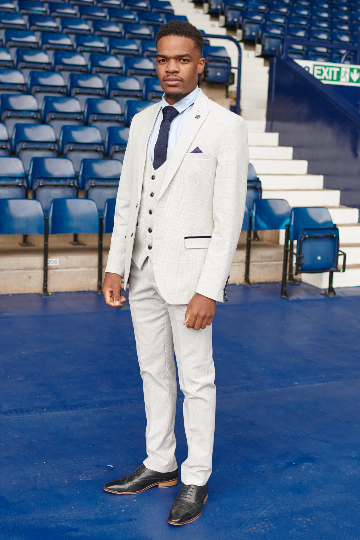 A Black man wears the Marc Darcy WBA Collection Bromley Suit as worn by Grady Diangana, featuring a light Prince of Wales check slim fit, while standing in front of blue stadium seats.